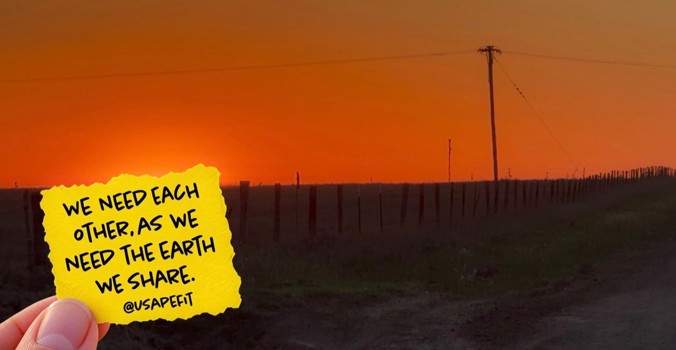 A hand holds a yellow sticky note at sunset reading 'We need each other, as we need the earth we share' by a rural road.