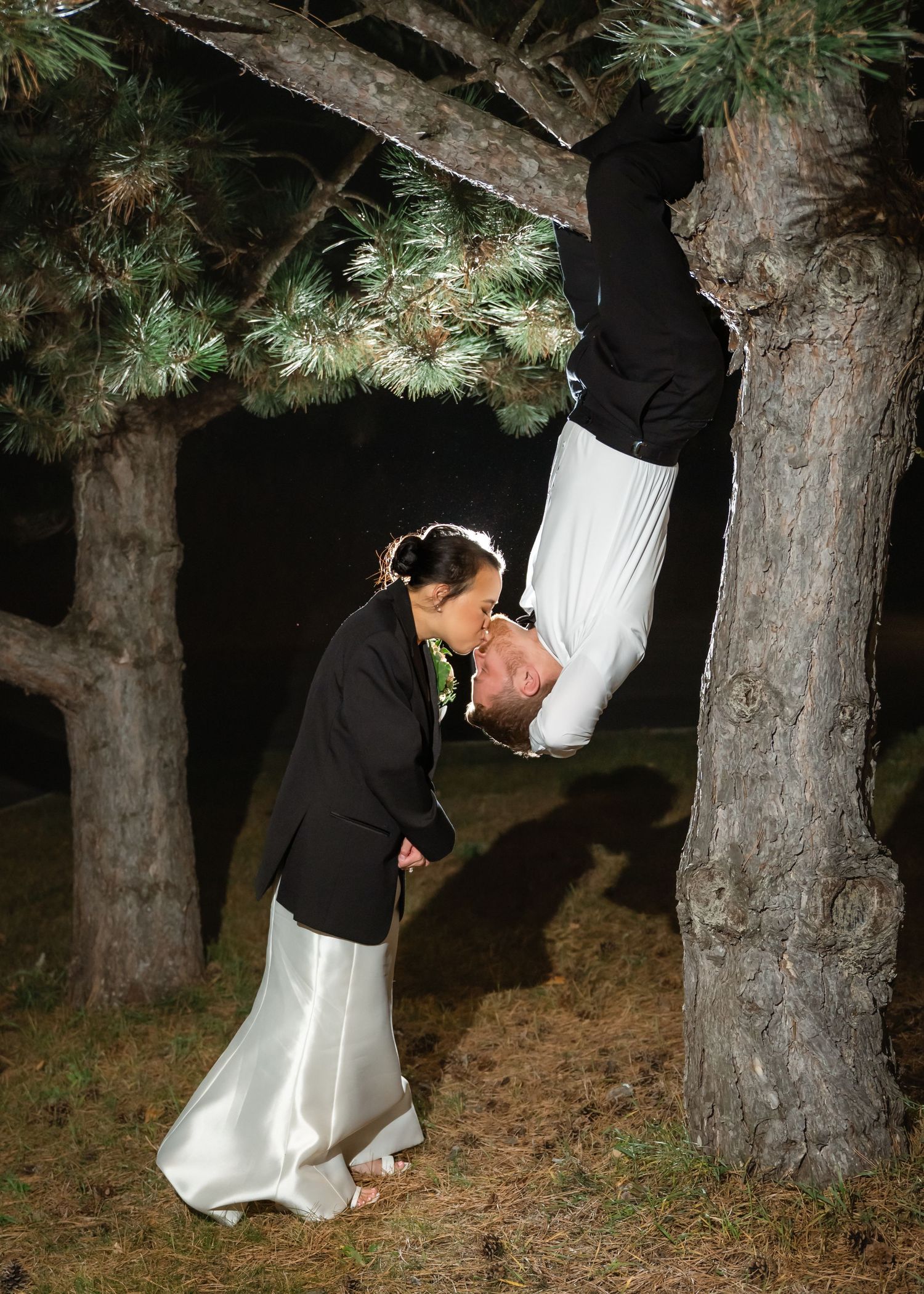 A bride in a white satin skirt and black jacket kisses a groom hanging upside down from a pine tree branch at night.
