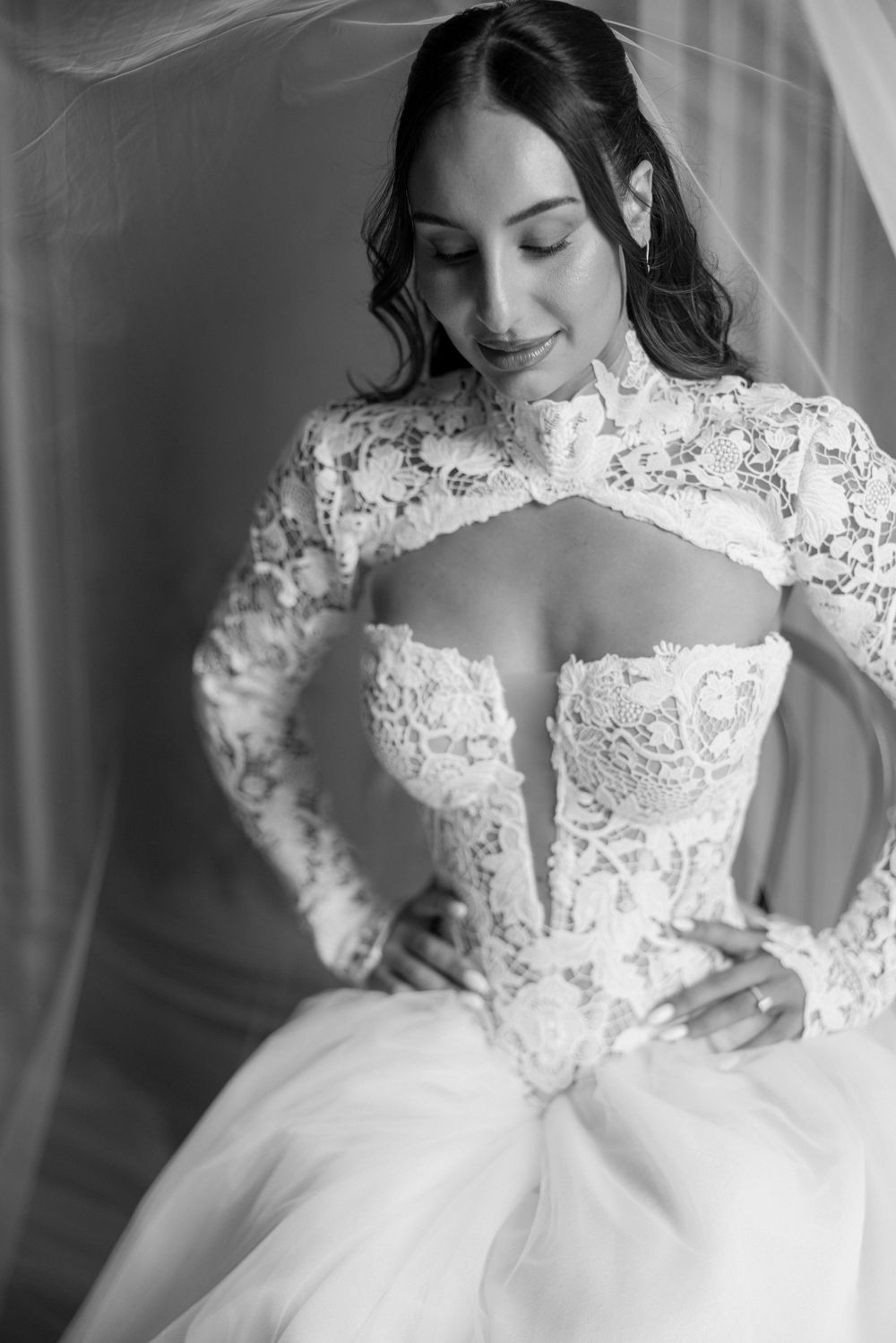 Black and white portrait of a bride in a long-sleeve lace bolero and corset bodice gown with a tulle skirt, hands on hips.