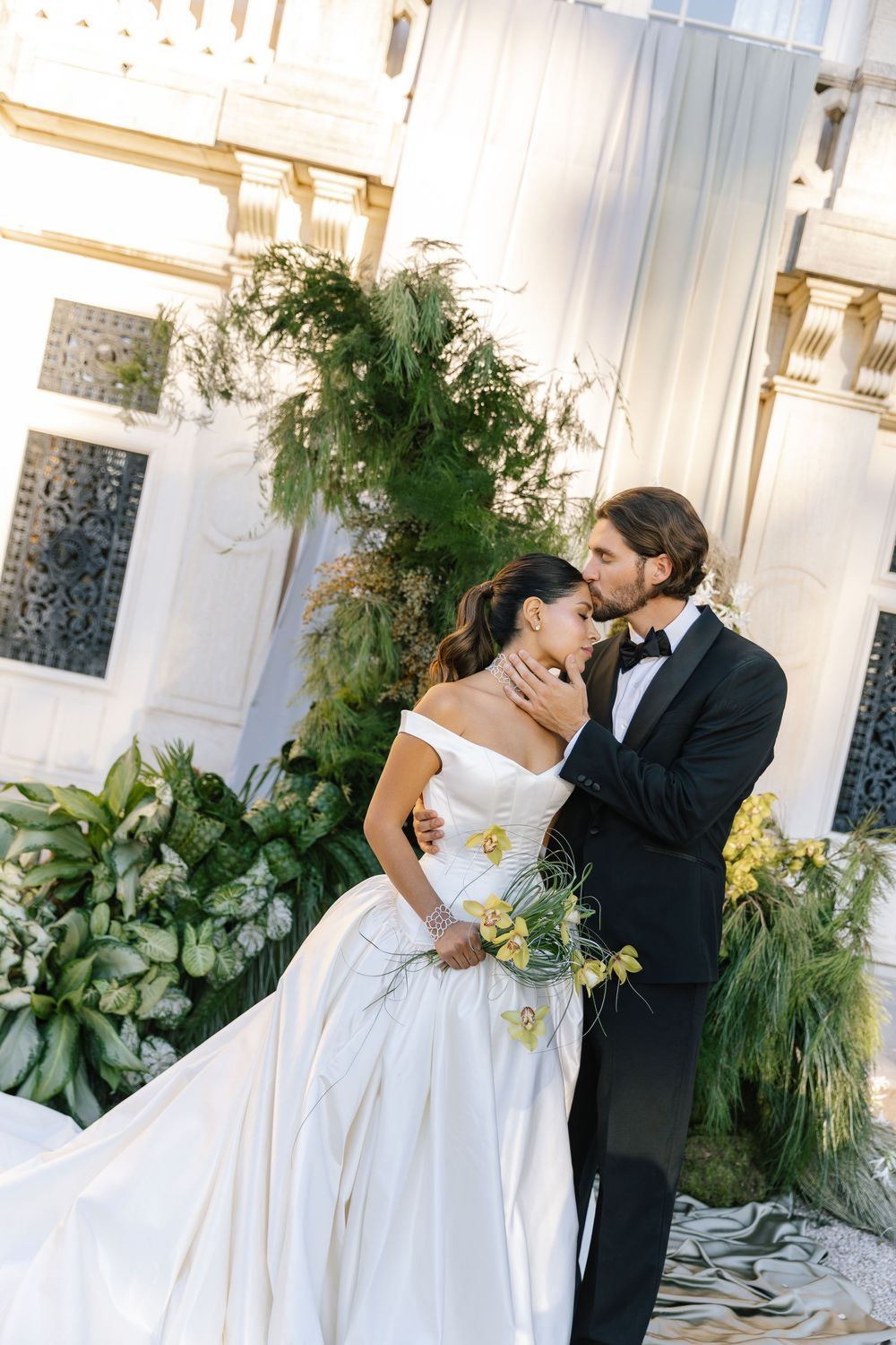 Groom in black tuxedo and bow tie kisses bride's forehead, her off-shoulder satin gown and yellow bouquet set against lush greenery.