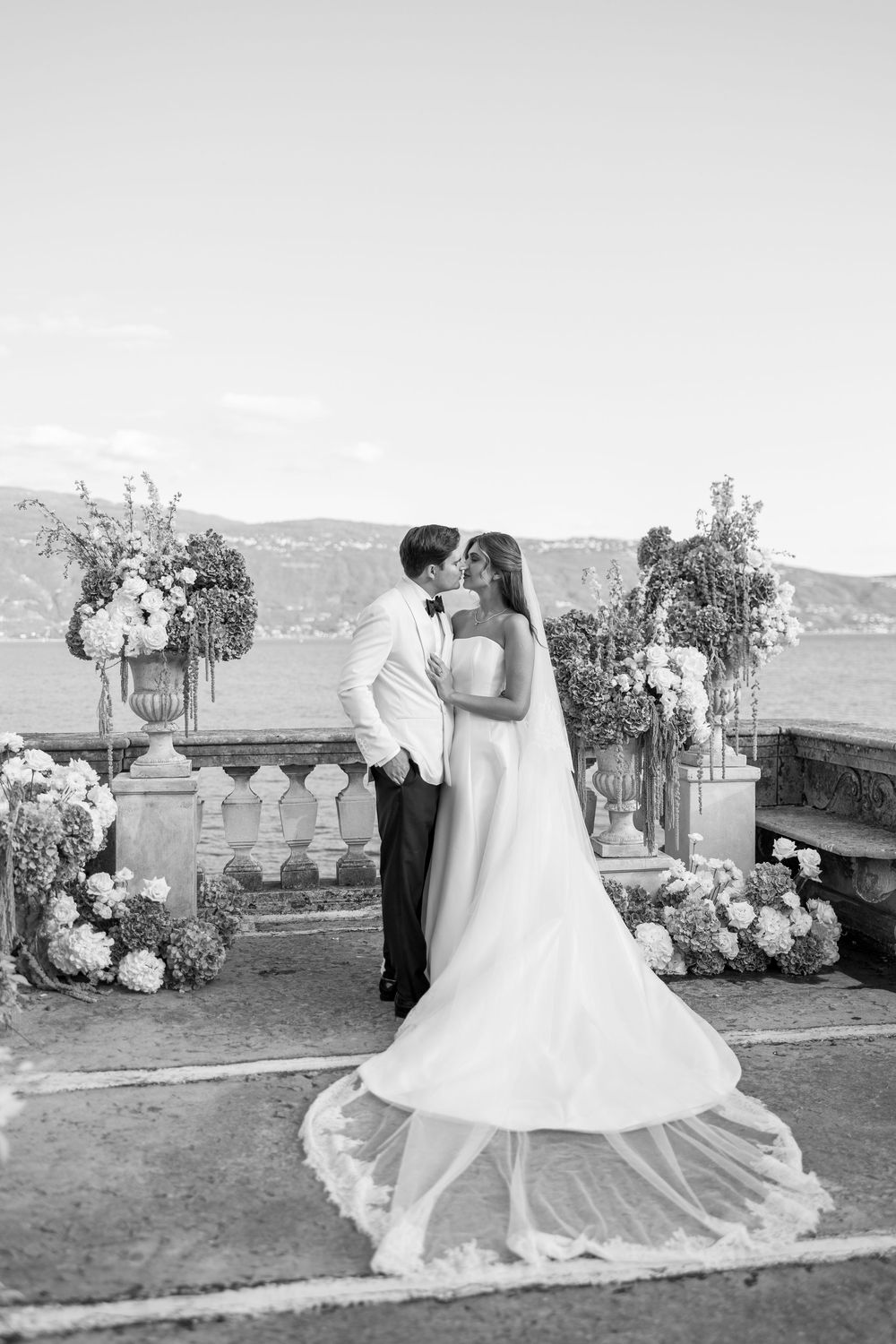 Black and white photo of a bride and groom kissing on a lakeside terrace with floral urns, a long veil and mountains behind.