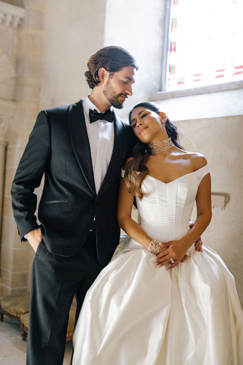 Bride in an off-shoulder satin wedding gown rests her head on the groom's chest as he wears a black tuxedo and bow tie.