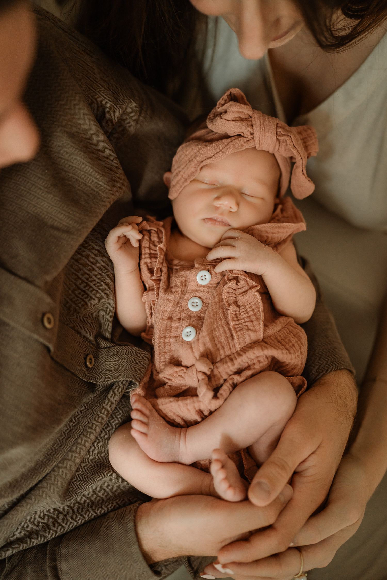 Sleeping newborn baby in a dusty pink muslin romper and bow headband, cradled in the arms of two parents during a cozy family photo session.