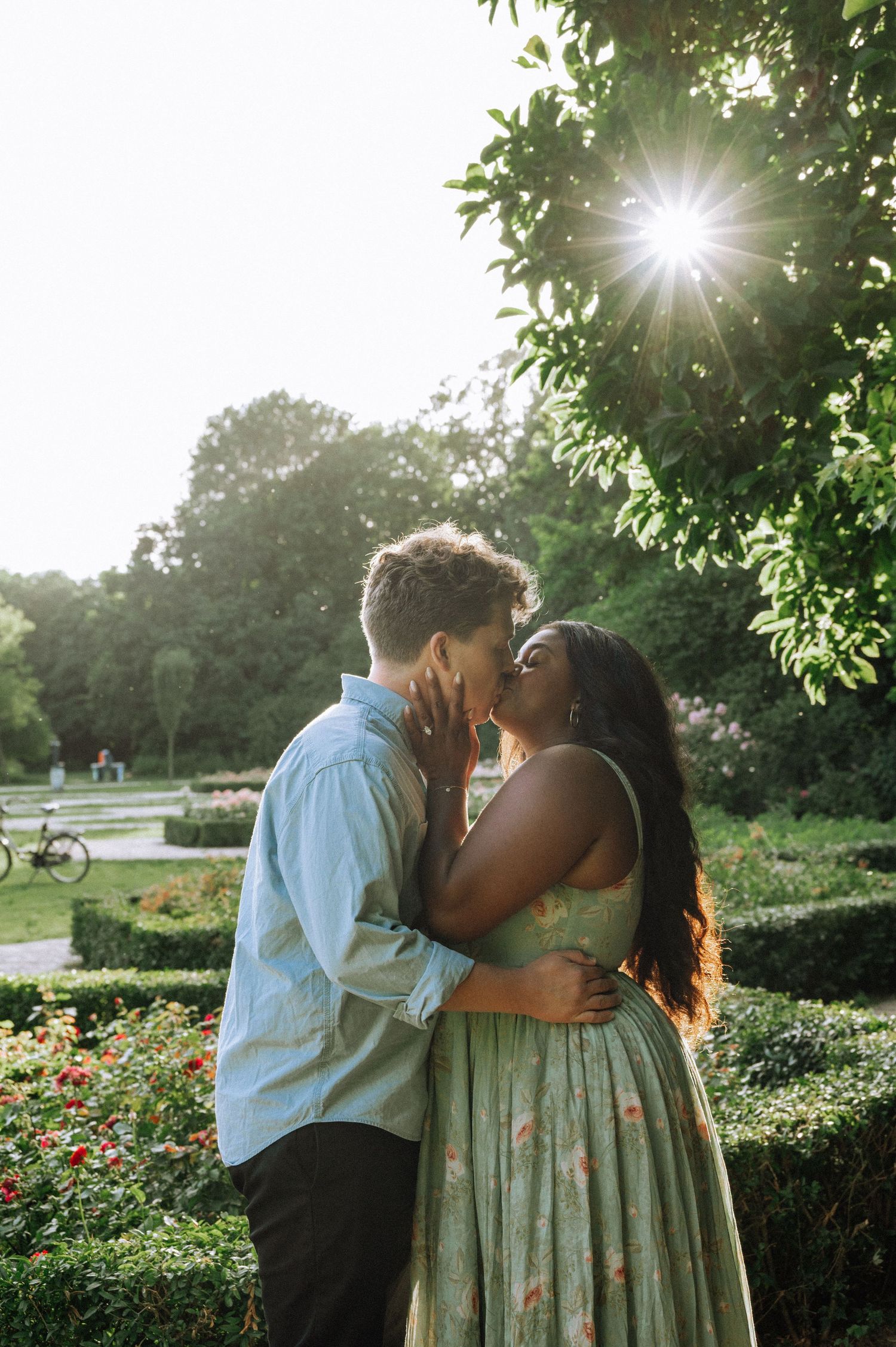 Couple in light summer clothing share a romantic kiss in a sunlit garden with green hedges, red flowers and trees in Vondelpark, Amsterdam.