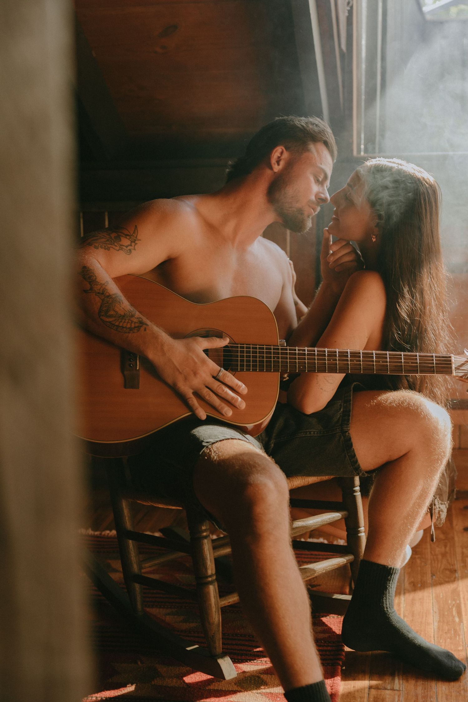 Shirtless tattooed man holding an acoustic guitar leans in for a kiss with a woman as sunlight streams through a rustic wooden cabin.