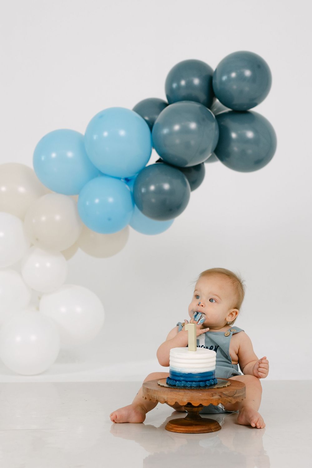 Baby in denim overalls sits on a white floor eating a small blue and white smash cake beside a blue, gray and white balloon garland.