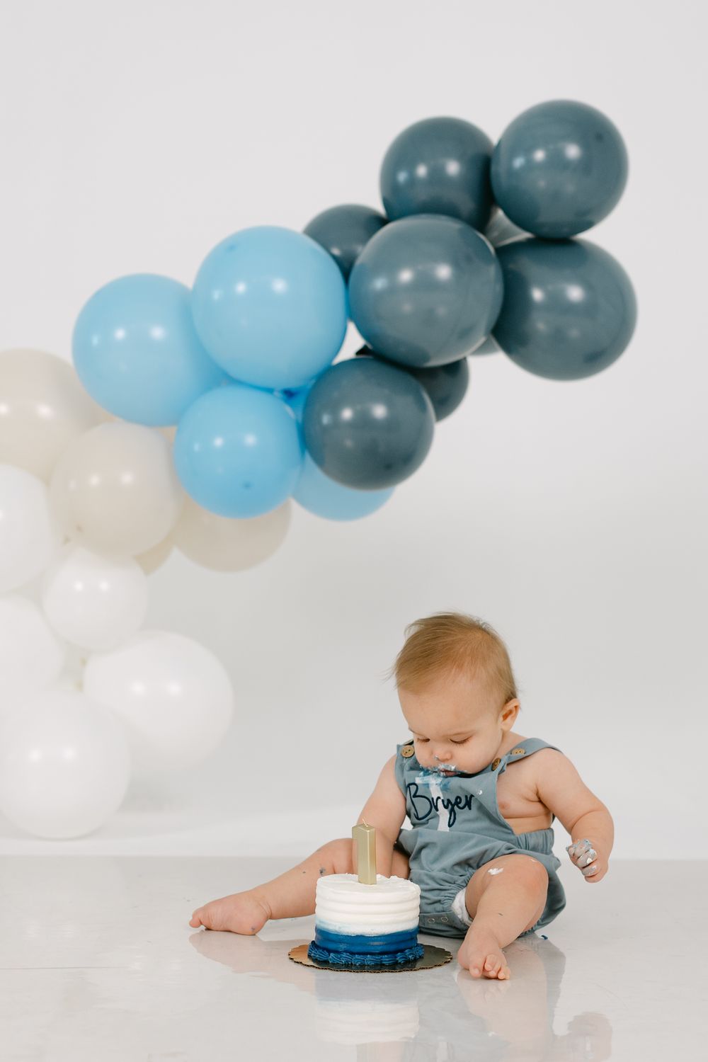 Baby in a blue romper sits on a white floor beside a small blue and white smash cake, with blue, gray and white balloons above.