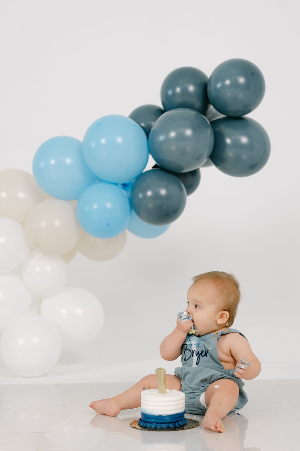 Baby boy in a denim romper eating a small blue smash cake beside a balloon garland of blue, slate and white balloons.