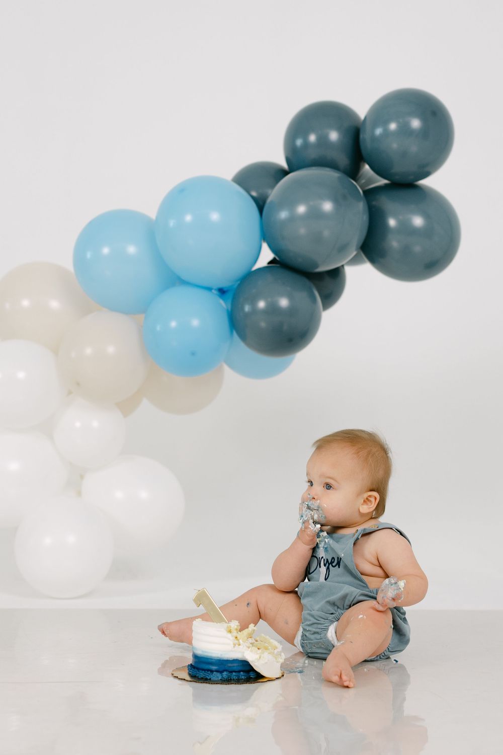 Baby in a gray romper sits on a white floor eating a blue smash cake beside a balloon garland of white, light blue and slate gray balloons.