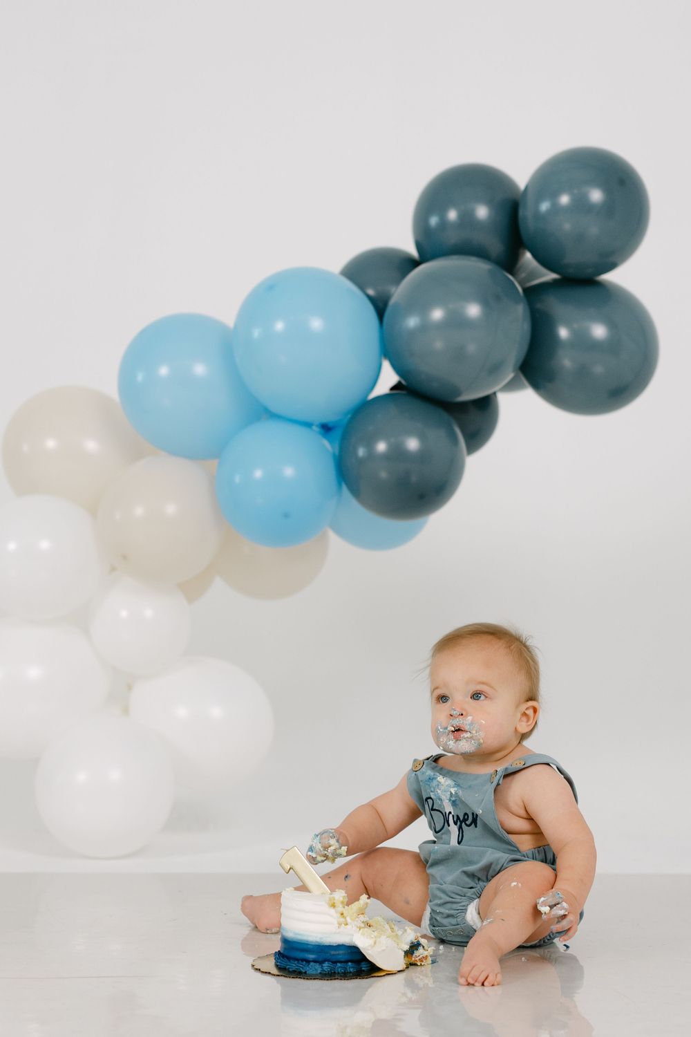 Baby in a gray romper sits on a white floor with frosting on his face, enjoying a smash cake beneath blue, gray and white balloons.