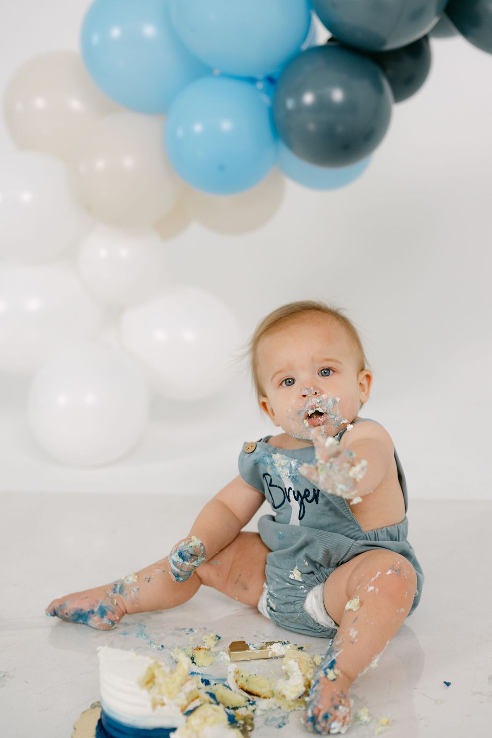 Messy baby in a gray romper with a name on it sits smiling beside a smashed cake with blue, gray and white balloons behind.