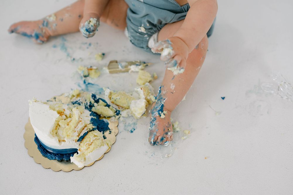 Messy baby in a gray outfit sits on a white floor, hands and legs covered in frosting after smashing a blue and white cake.
