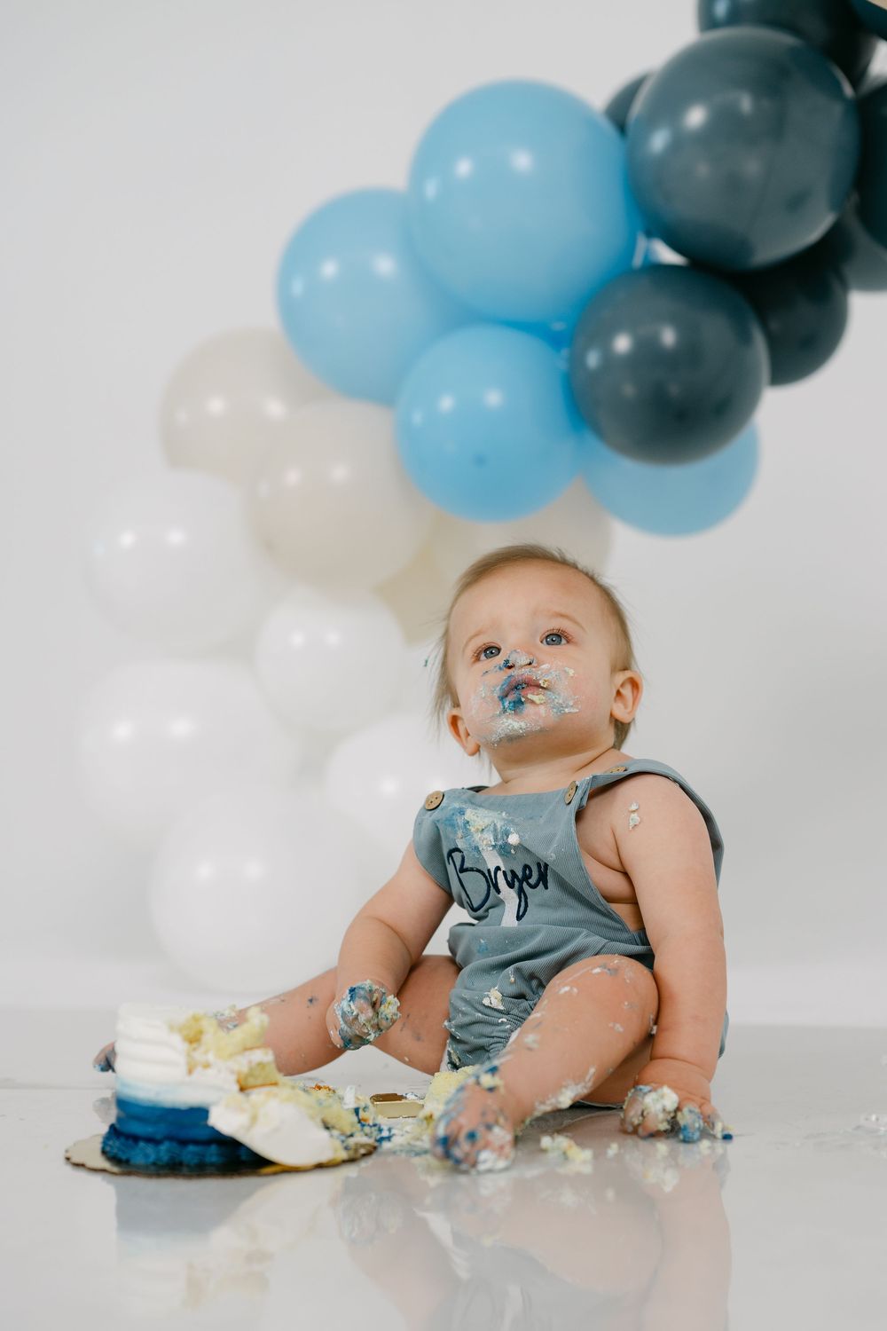 Baby in a gray romper sits on the floor covered in frosting during a first birthday cake smash, with blue, gray and white balloons behind.
