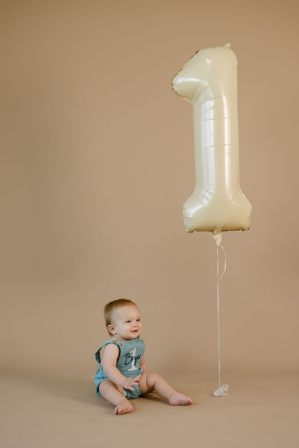 Baby in a blue romper sits smiling on a tan backdrop beside a cream number one foil balloon for a first birthday photo.