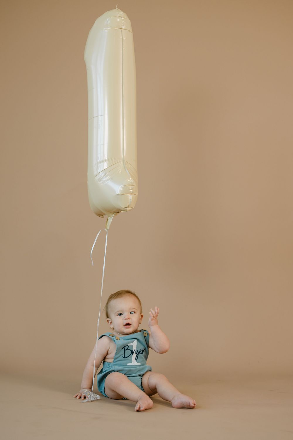 Baby in a teal outfit with a name on it sits barefoot and waves beside a cream number one balloon against a tan studio backdrop.