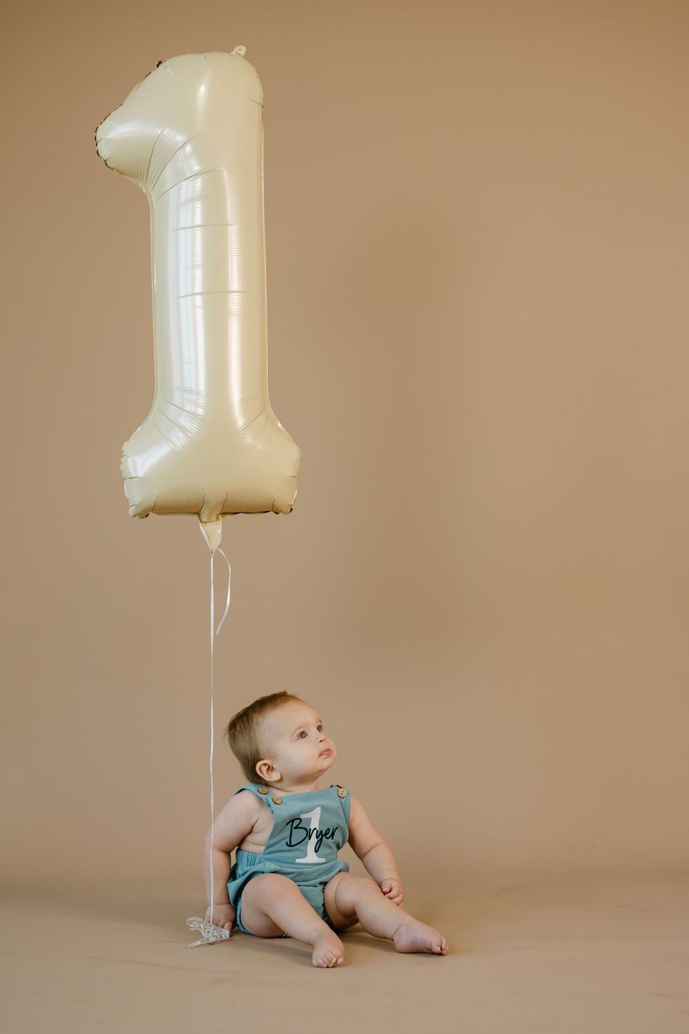 Baby in a teal romper sits on a tan backdrop gazing up at a large cream number one foil balloon for a first birthday photo.