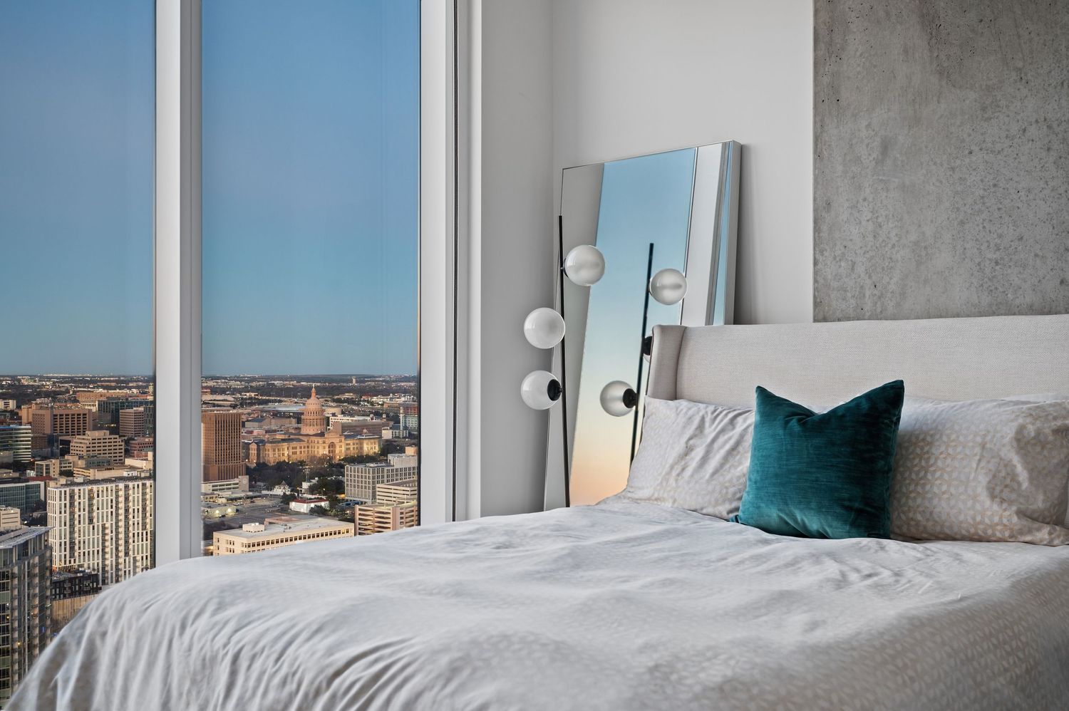 Modern bedroom with cityscape views, floor mirror, and teal accent pillow on white bedding.