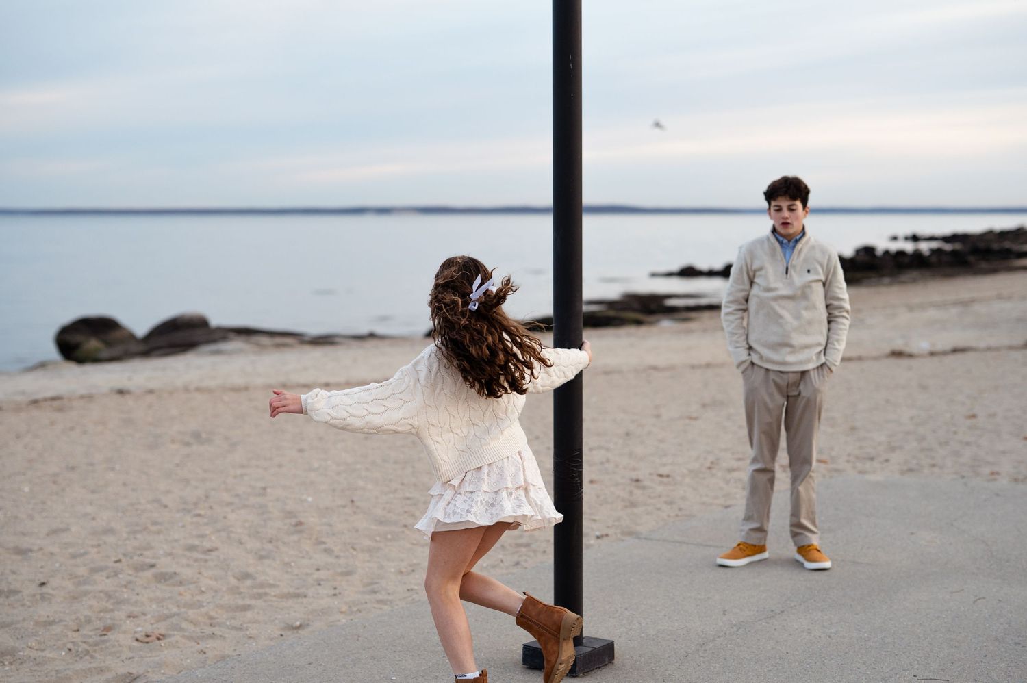 Siblings in cream sweaters smile together on a sandy beach in Connecticut with calm waters behind them.