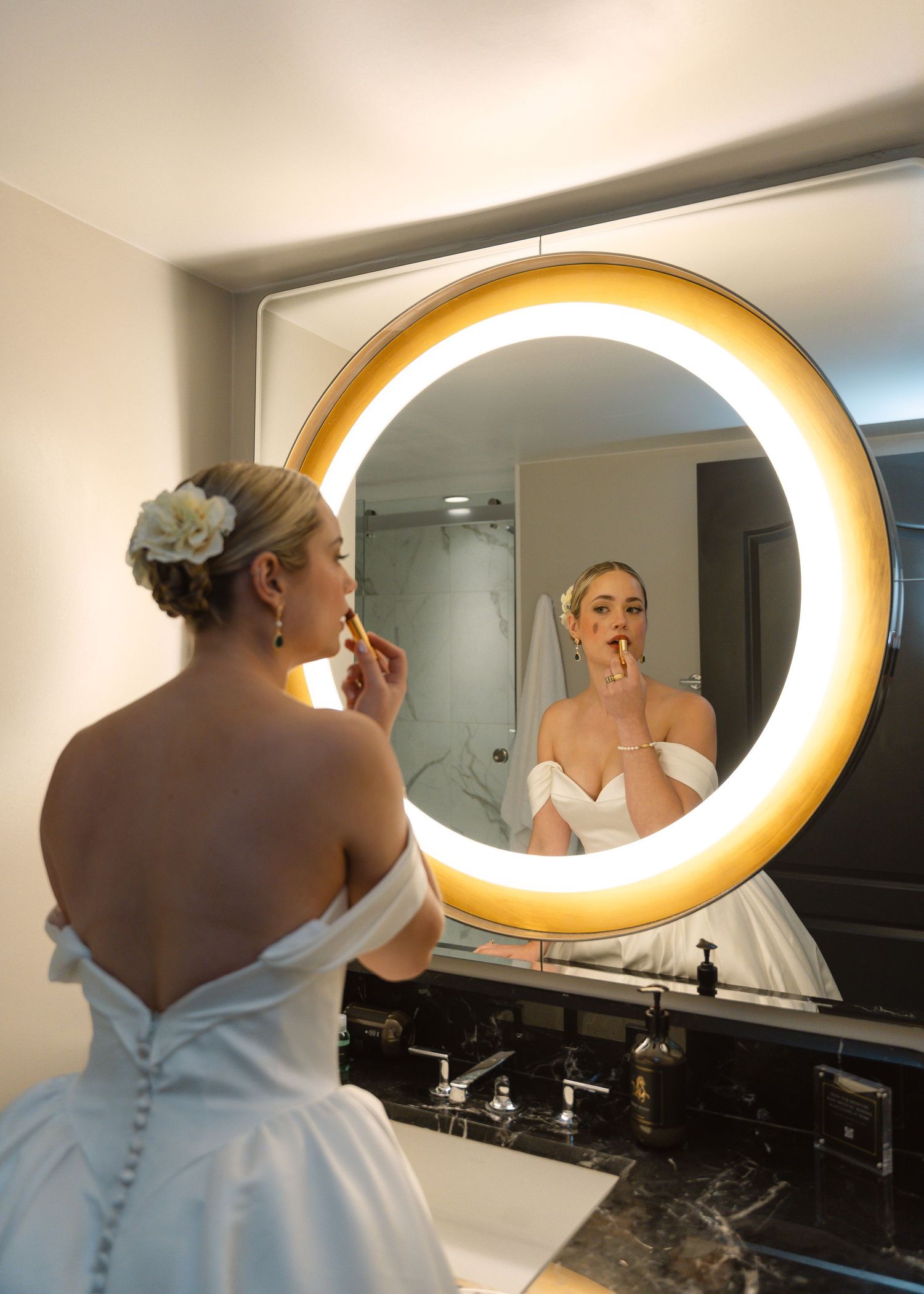 Bride in white off-shoulder gown applies makeup at illuminated round mirror in elegant bathroom.
