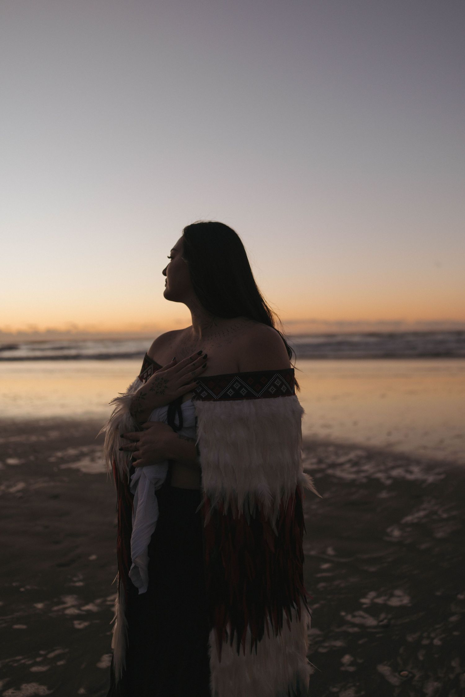 Woman in traditional Maori cloak stands on beach at sunset, hand on chest, silhouetted against golden sky.