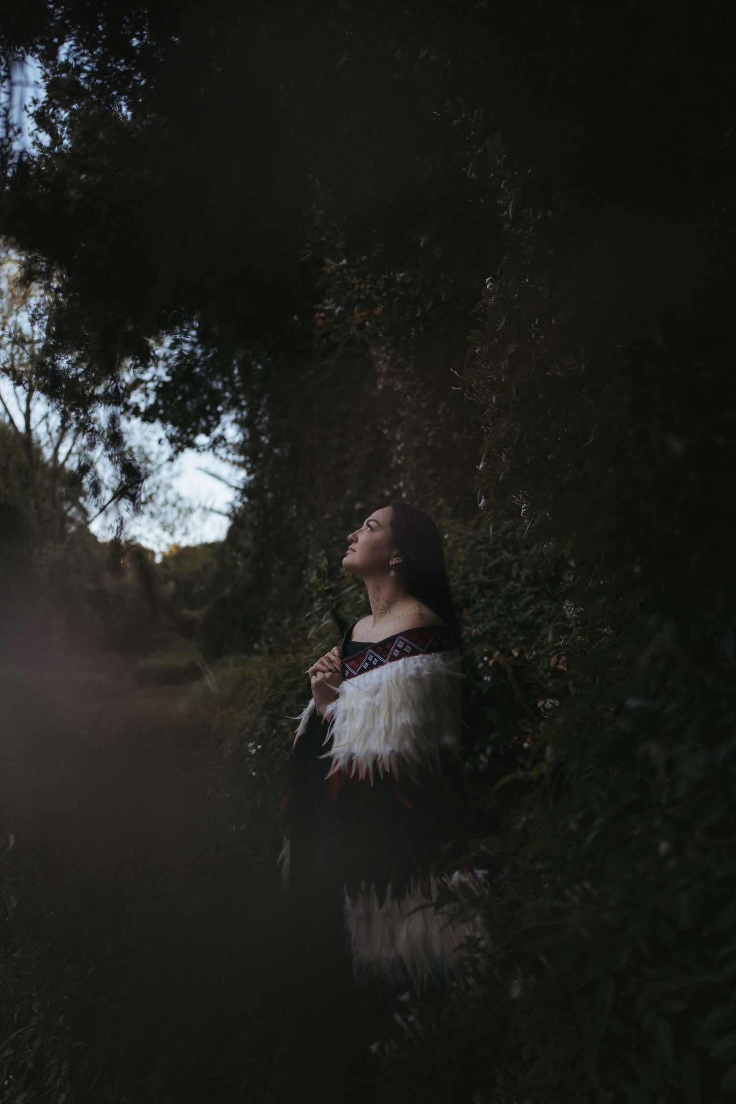 Woman in traditional Maori attire with feather cloak stands among lush green foliage in a dark, moody forest setting.