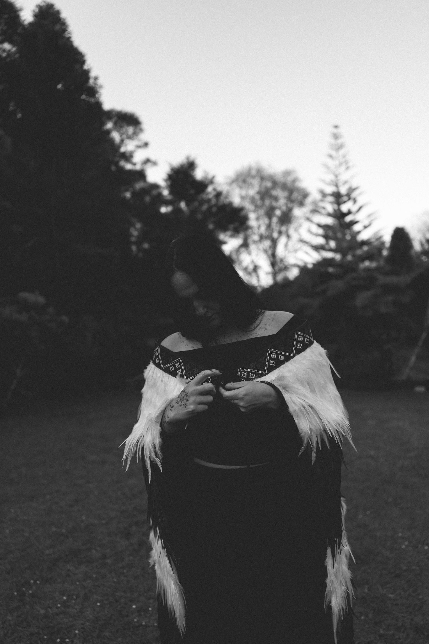 Black and white photo of a person wearing a traditional Maori cloak with feathers and geometric patterns outdoors.