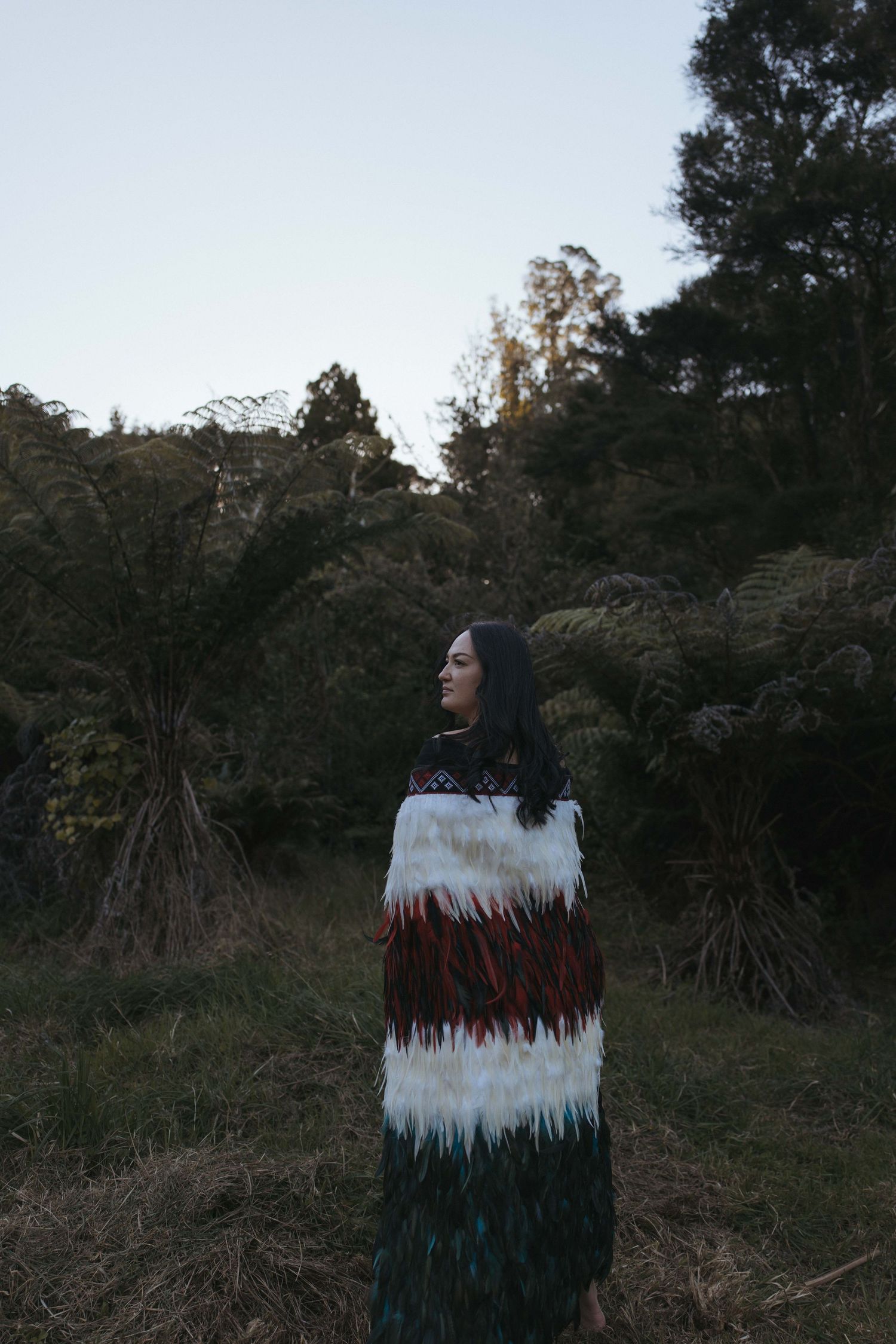 Māori woman wearing traditional feathered kākahu cloak stands in native New Zealand bush at dusk.