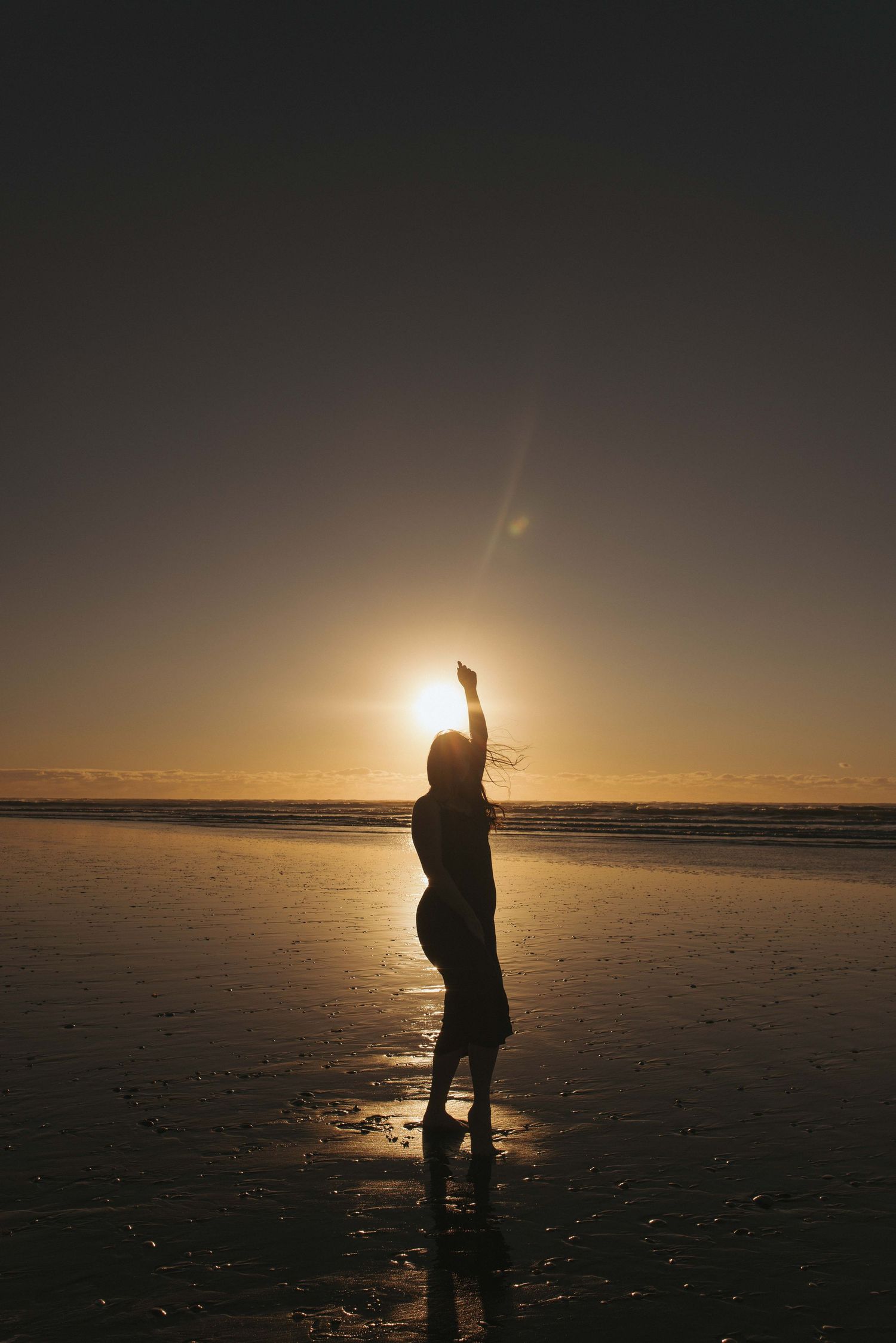 Silhouette of a person raising their arm toward the golden sunset on a reflective beach at low tide.