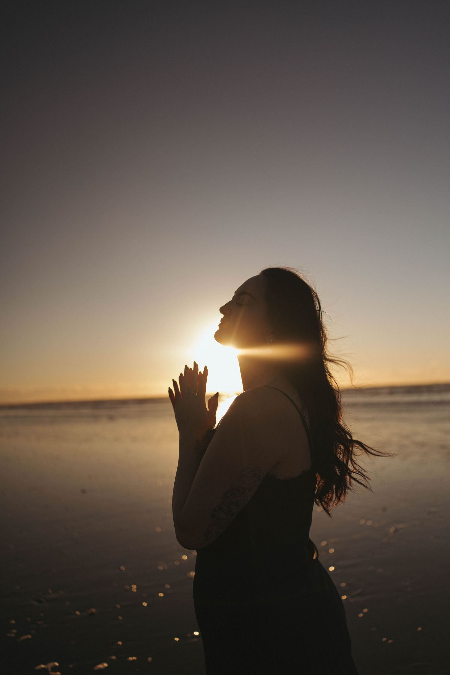 Silhouette of a woman praying at sunset on a beach, with golden light glowing behind her and hair blowing in the wind.