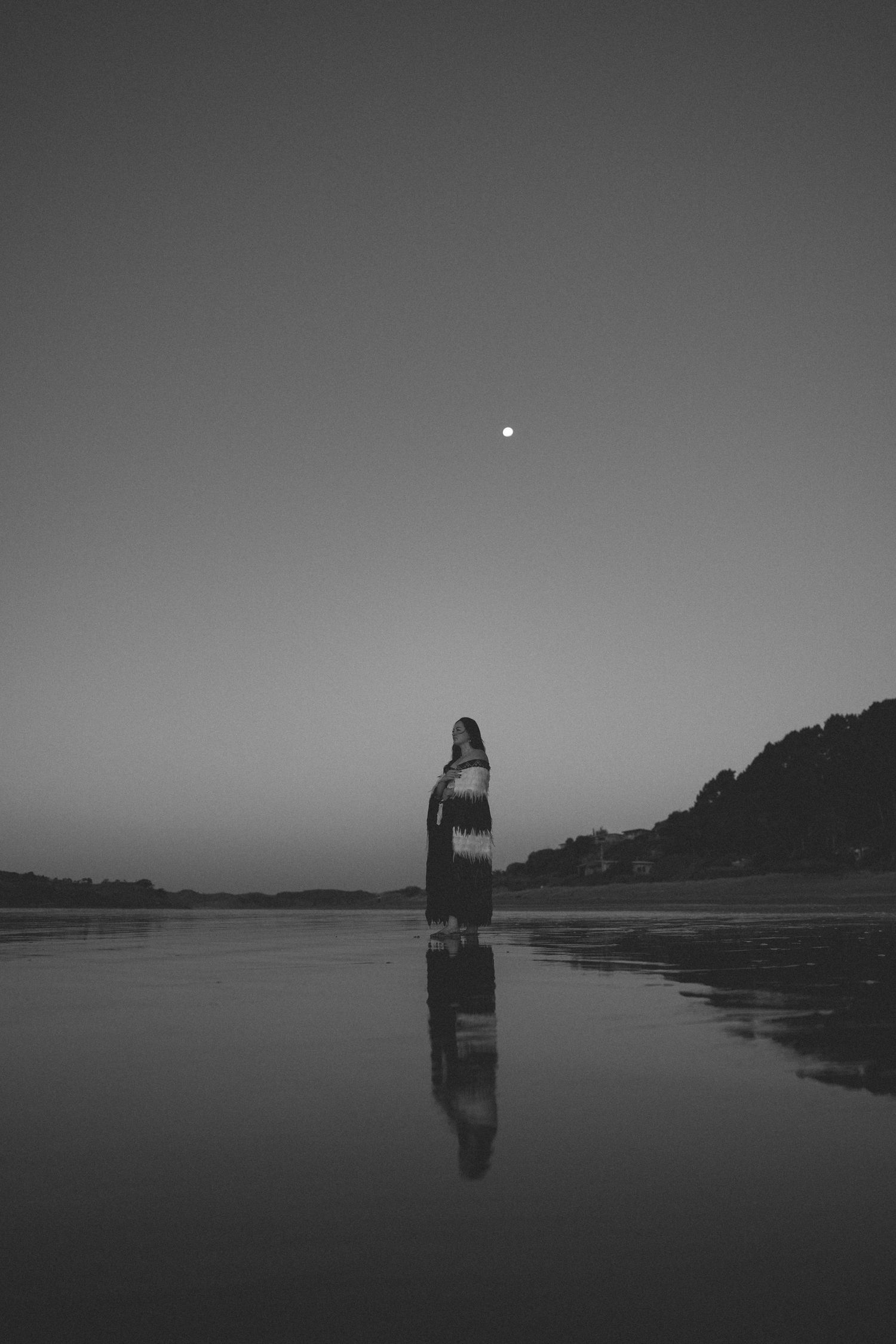 Black and white photo of a woman standing on a reflective beach at dusk with a full moon in the sky.