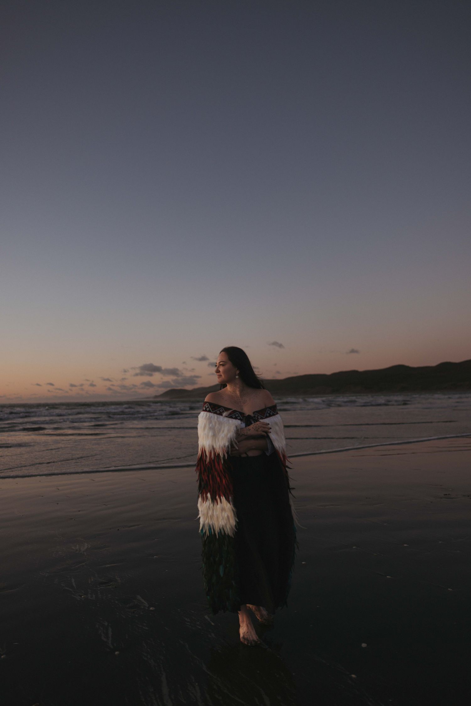 Woman in traditional Maori cloak stands barefoot on a beach at sunset, ocean waves behind her.