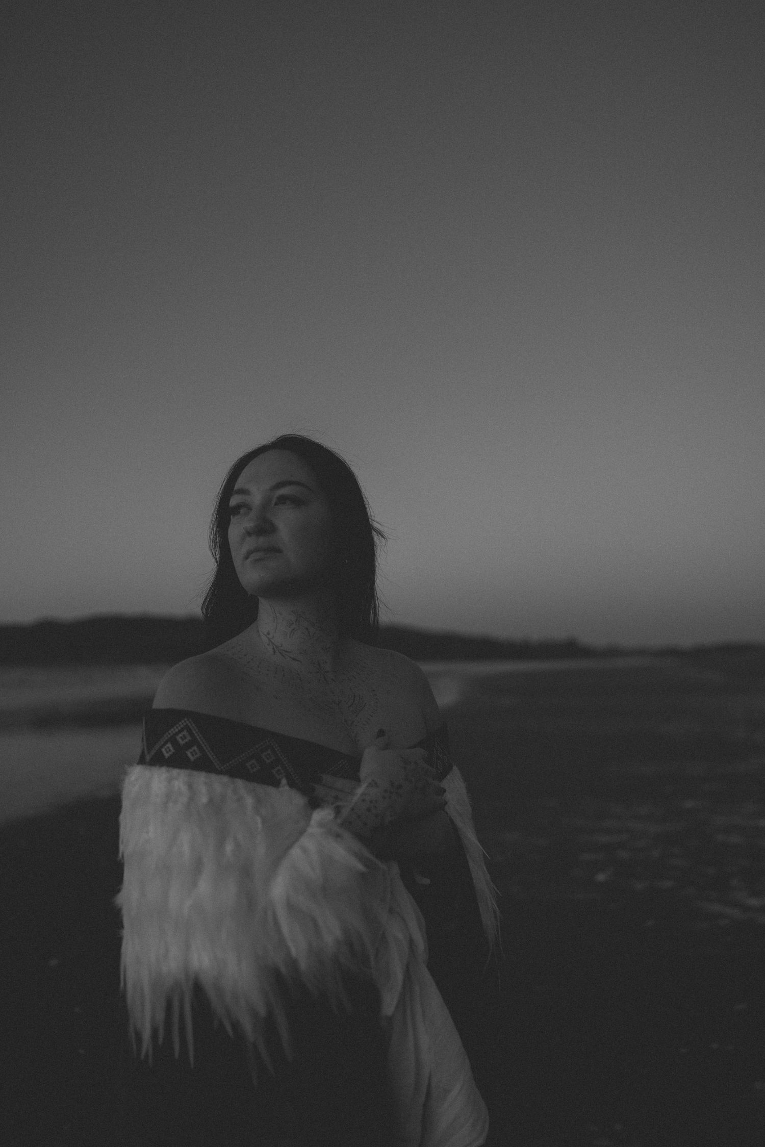 Black and white photo of a woman in a feathered garment standing outdoors at dusk, gazing upward thoughtfully.