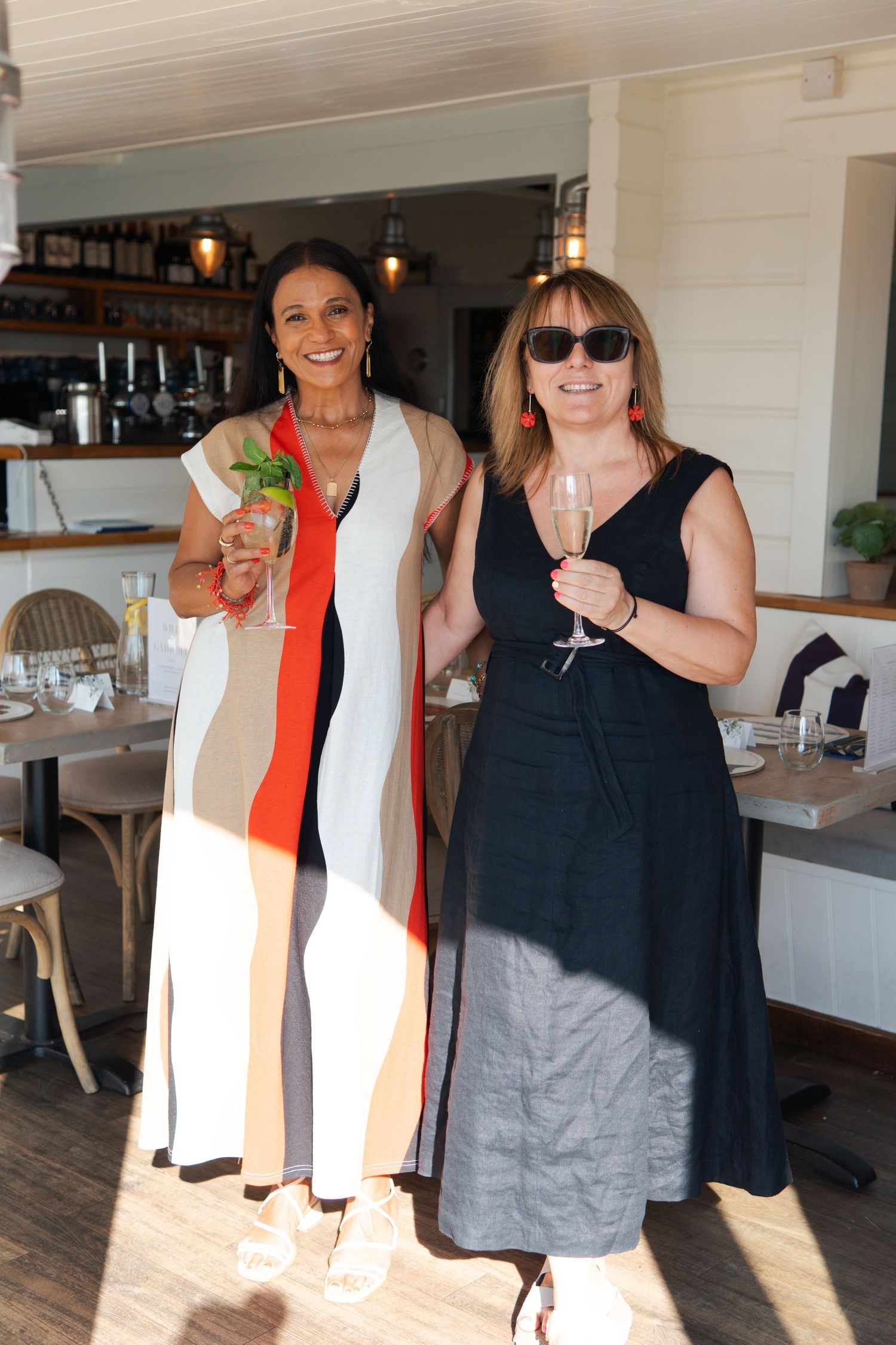 Woman in sunglasses and green top smiling at a table covered with magazines and wine glasses indoors.