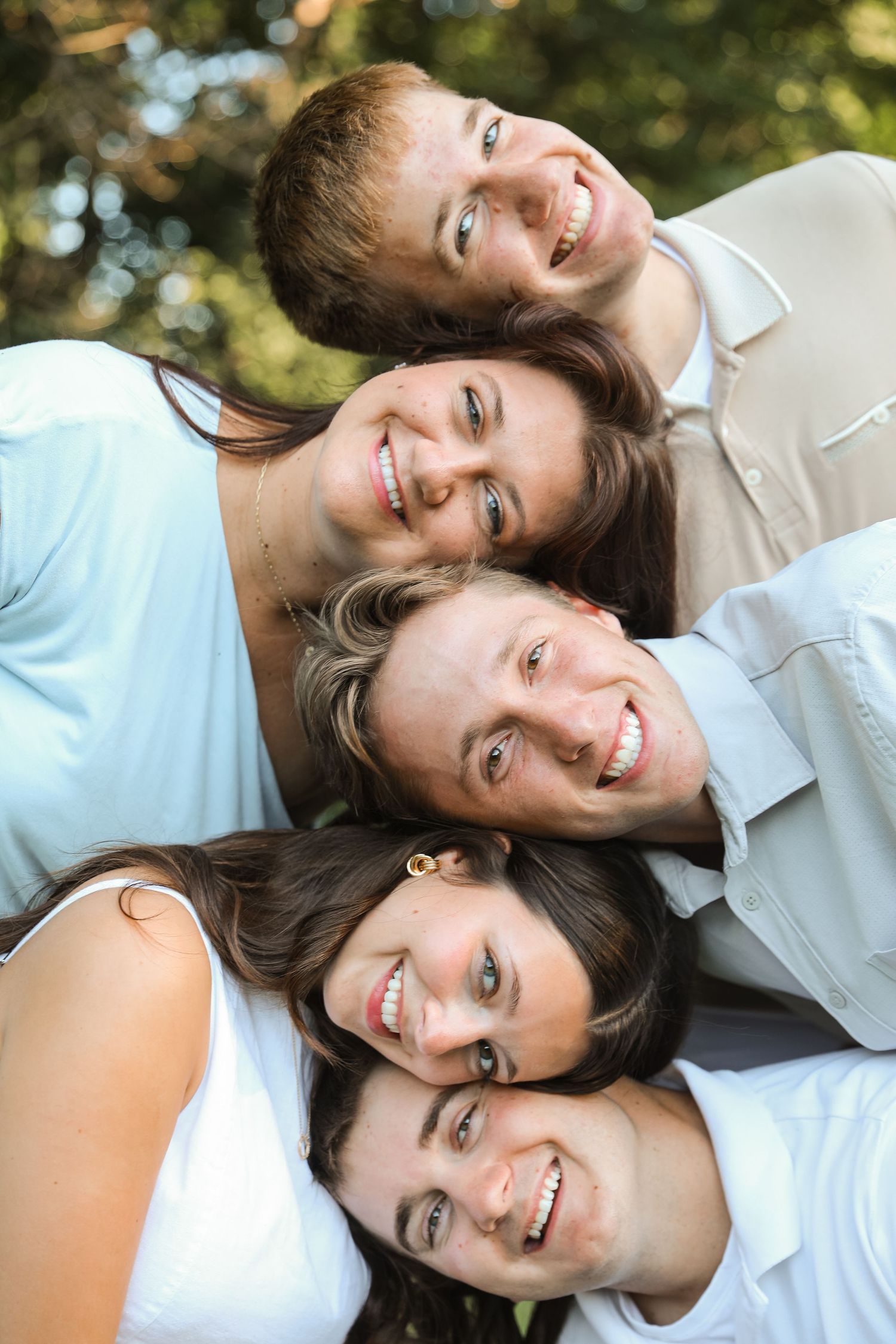 Five smiling friends pose together outdoors, stacking their heads in a fun vertical arrangement with green trees behind.