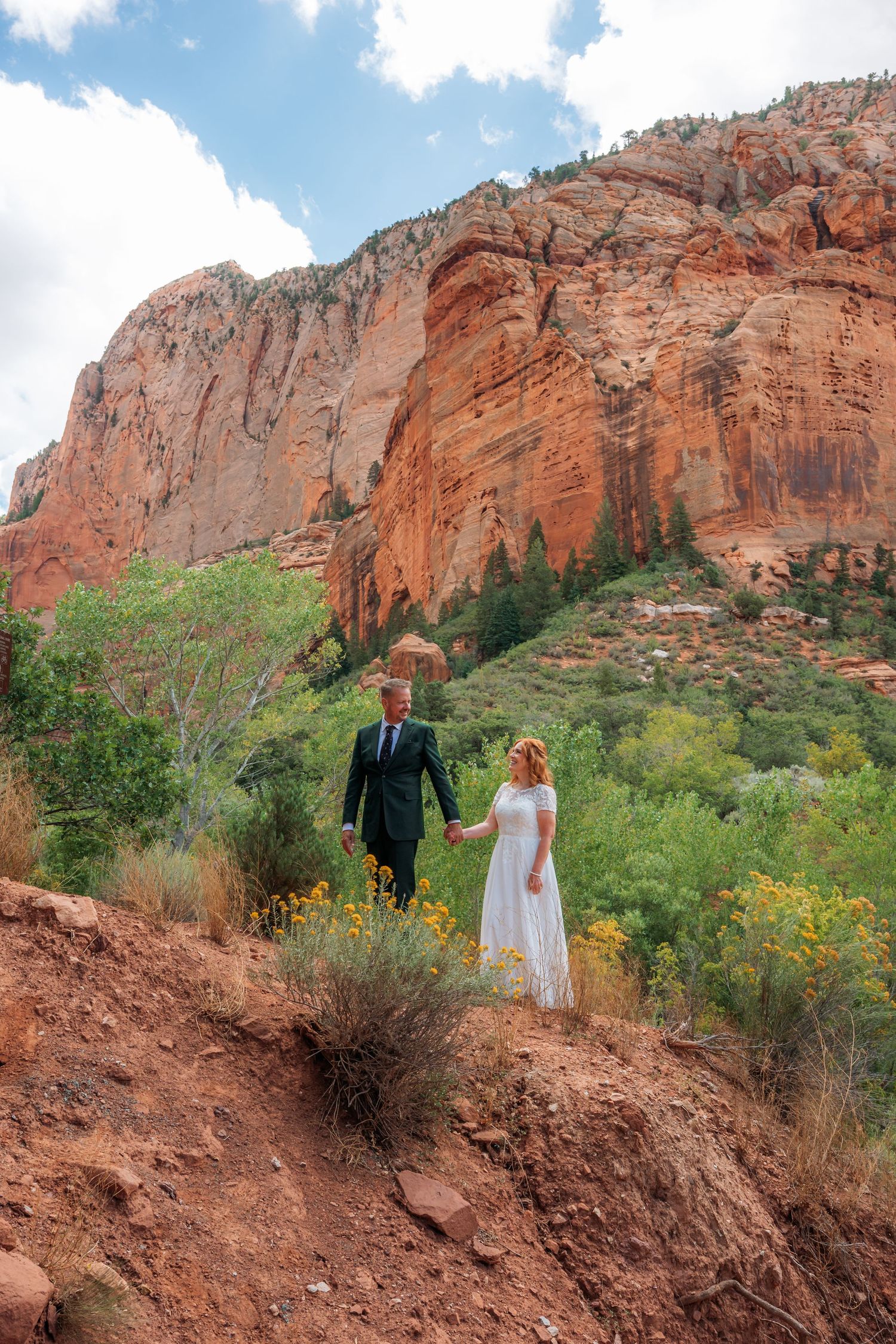 Bride and groom hold hands on a red rock trail with lush greenery and towering sandstone cliffs in Sedona, Arizona.
