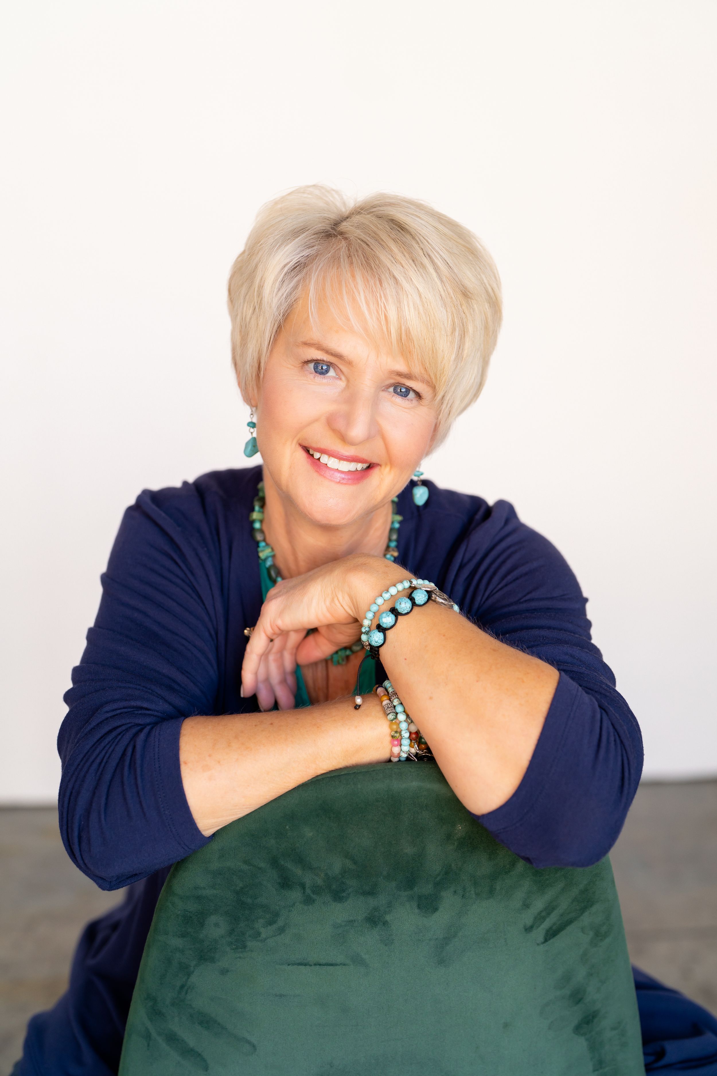 Smiling middle-aged blonde woman in navy blue top with turquoise jewelry, leaning on green velvet chair.