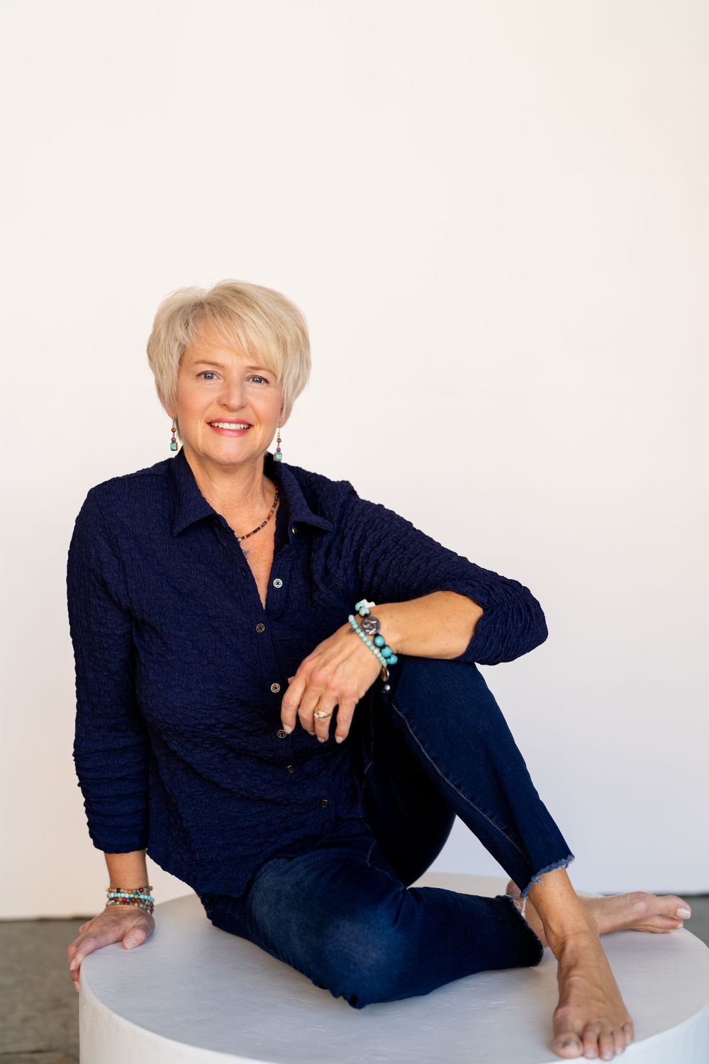 Smiling blonde woman in navy shirt and jeans sitting cross-legged on white surface wearing turquoise jewelry.