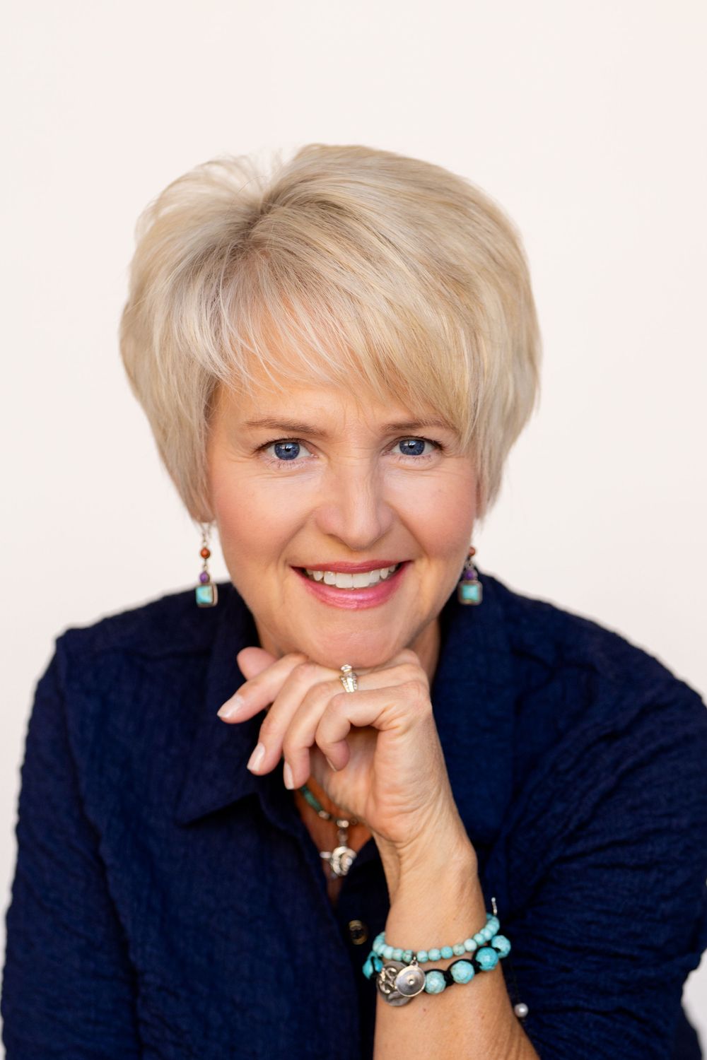 Smiling blonde woman with turquoise jewelry and navy blazer poses with hands folded, professional headshot photo.