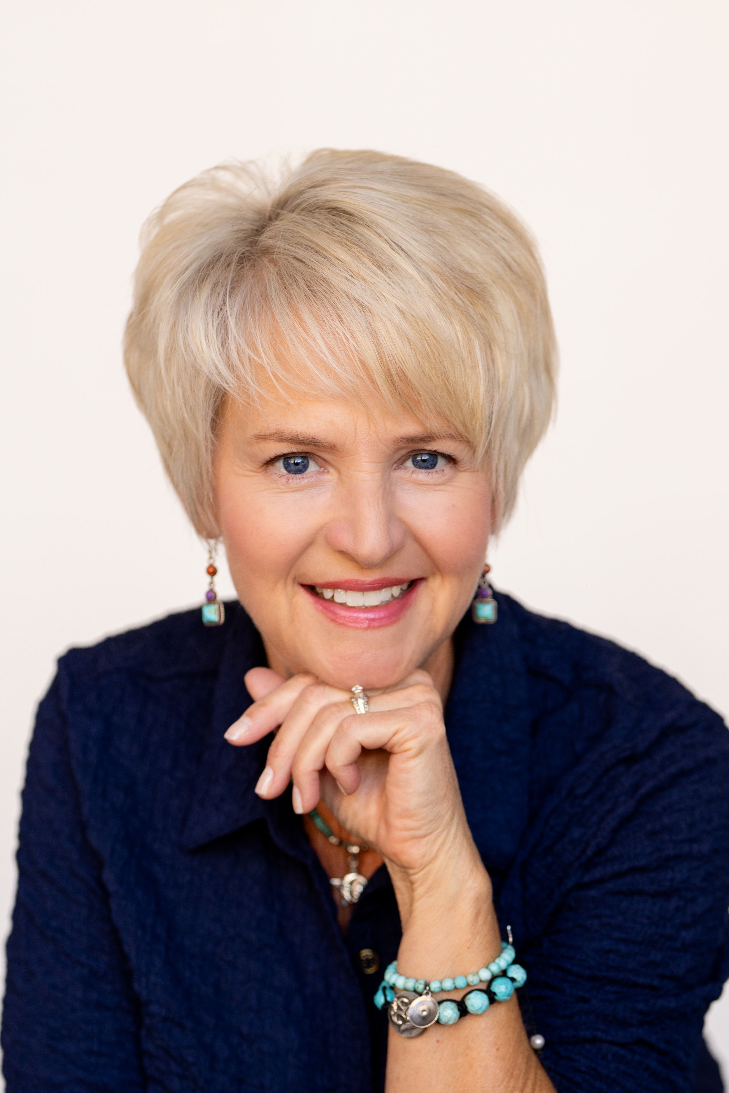 Smiling blonde woman with turquoise jewelry and navy blazer poses with hands folded, professional headshot photo.