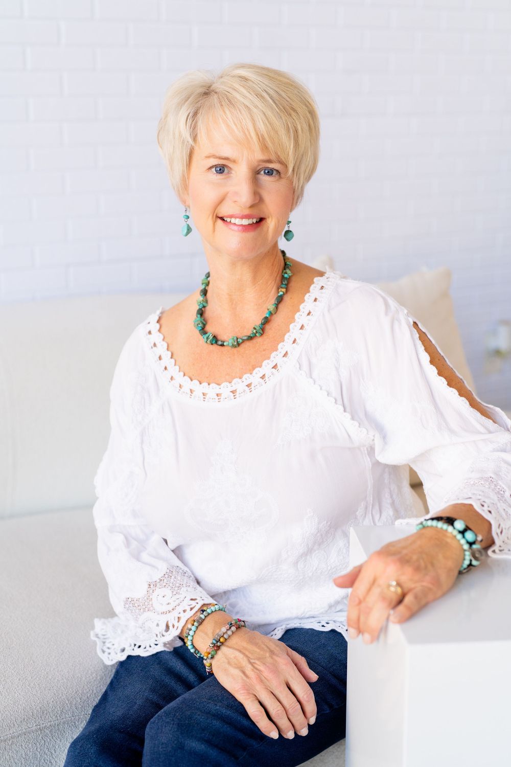 Smiling senior woman with short blonde hair wearing white blouse and turquoise jewelry seated on white sofa.