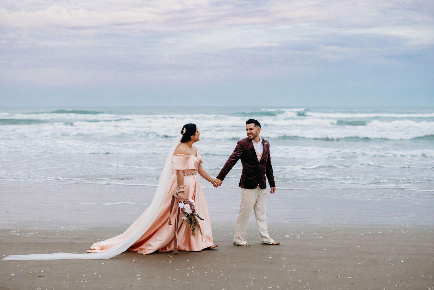 Bride in flowing pink gown and groom walking hand in hand on beach.
