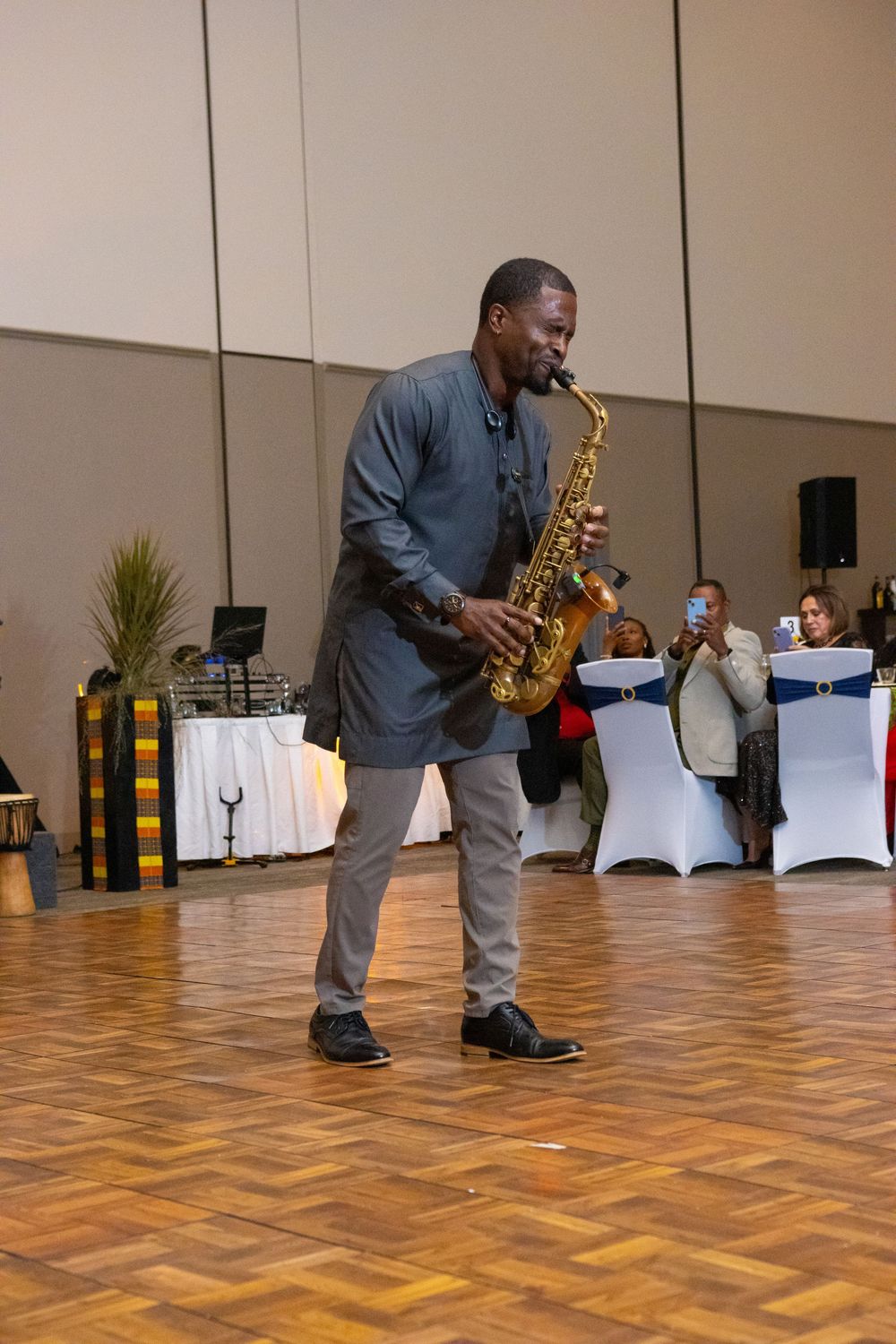 Man playing alto saxophone on a wooden dance floor at an indoor event with seated guests in background.