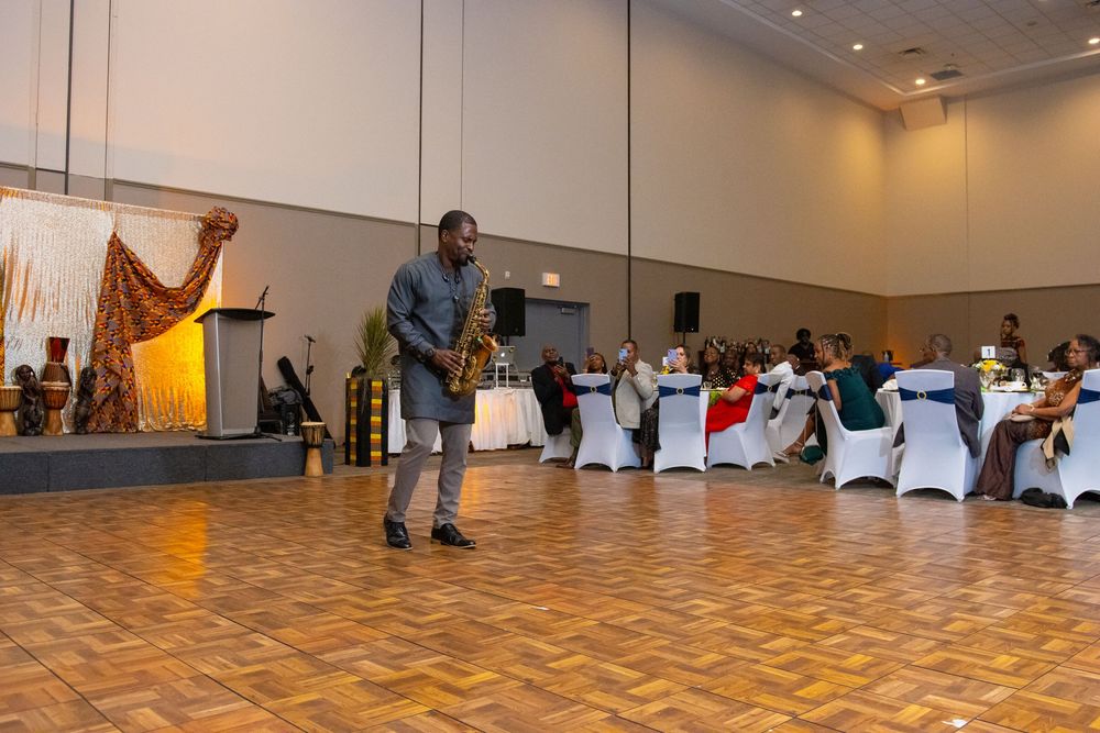 A saxophonist performs for guests at an elegant banquet hall event with white chair covers and decorative draping.