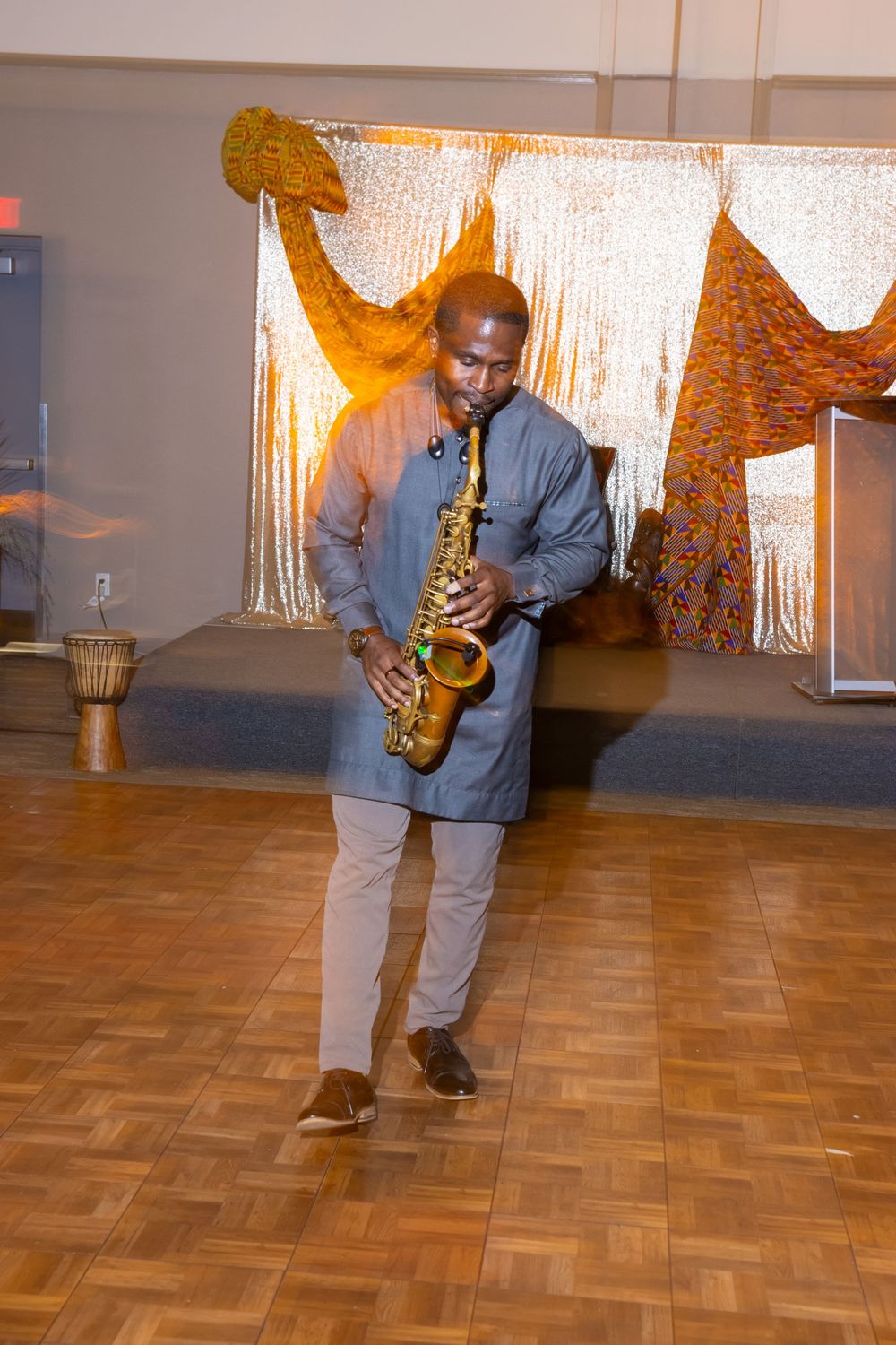 Man in gray outfit playing alto saxophone on wooden dance floor at an elegant event with decorative backdrop.