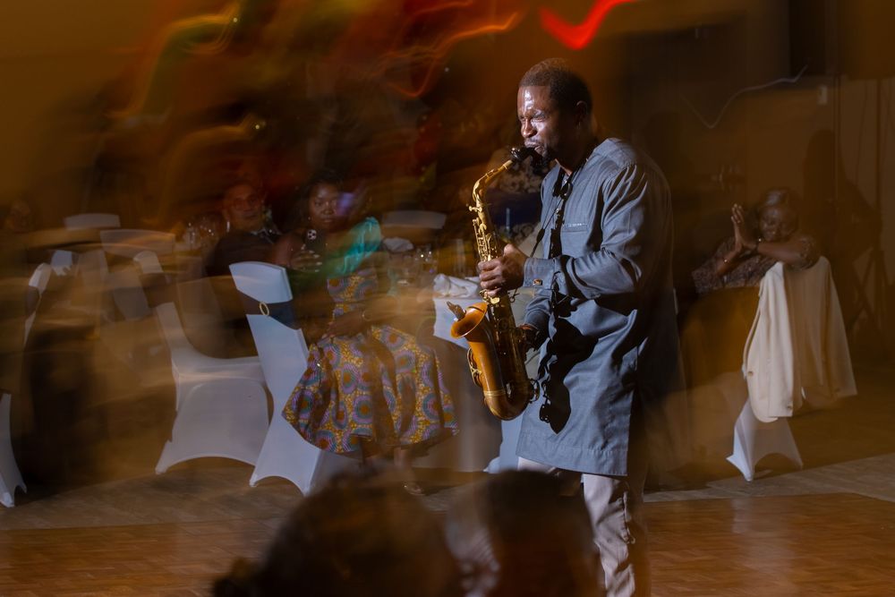 A saxophonist performs at an indoor event, with motion-blurred guests seated at white-draped tables in the background.