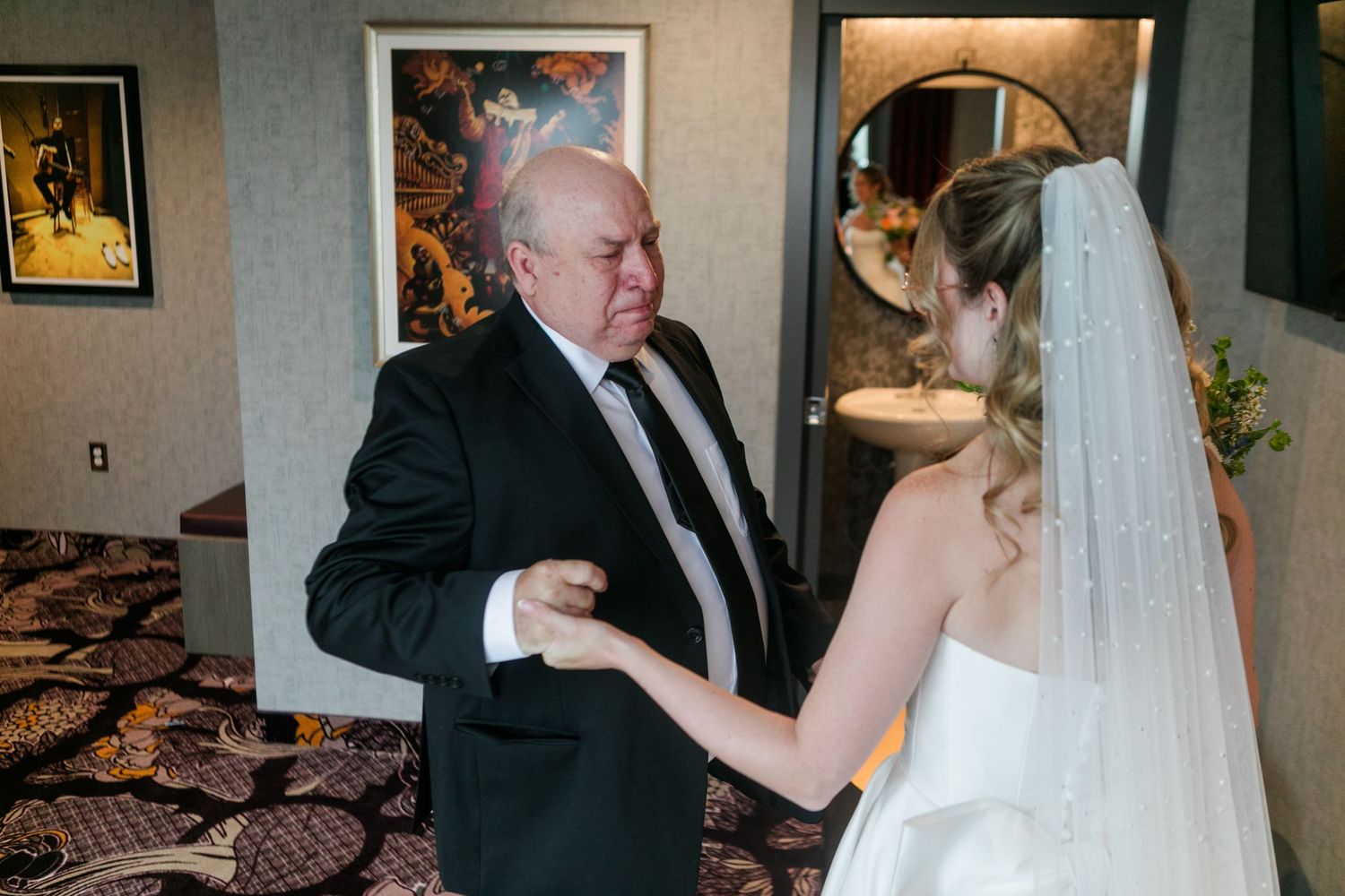 Emotional father and bride sharing a tender moment before wedding ceremony, man in black suit, bride in white gown with veil.