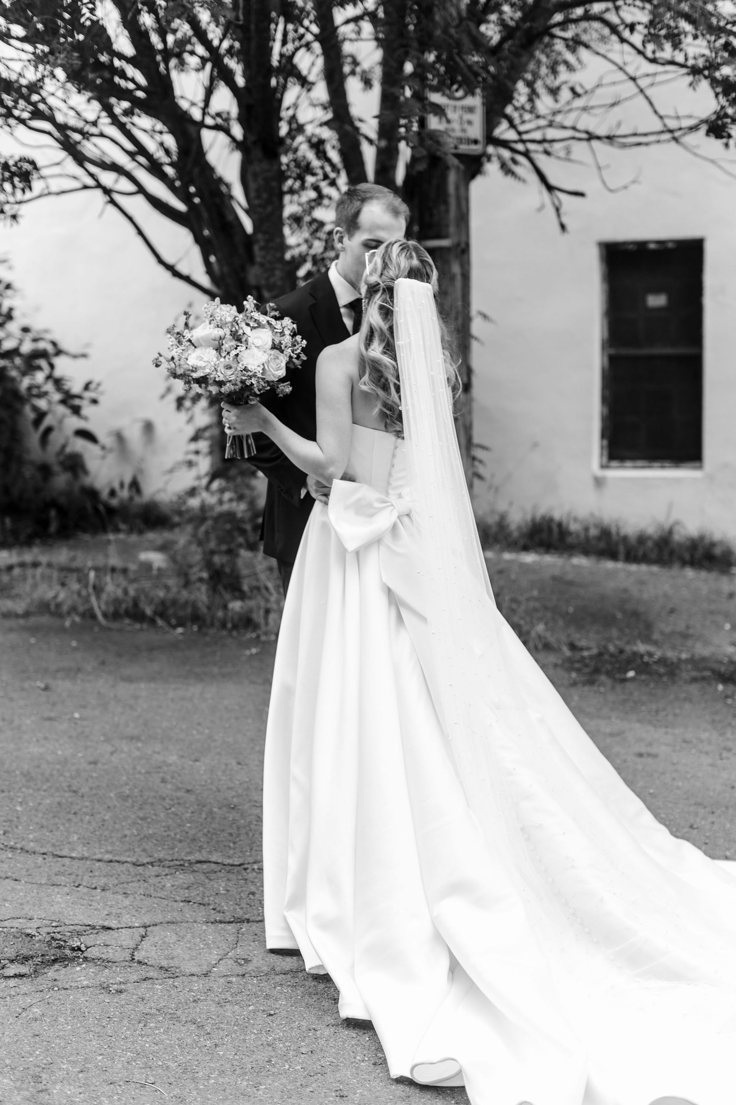 Black and white photo of a bride and groom kissing outdoors, her long veil trailing on the ground as she holds a bouquet.