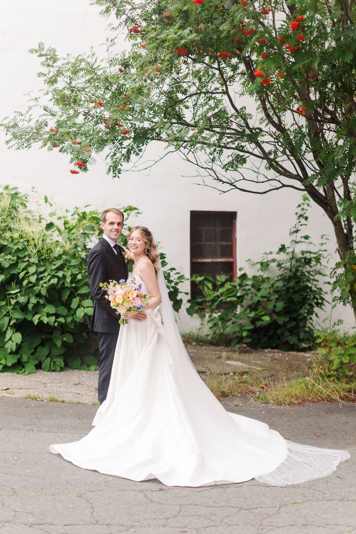 Bride and groom pose outdoors by white building with lush greenery and red berries, bride holding colorful bouquet.
