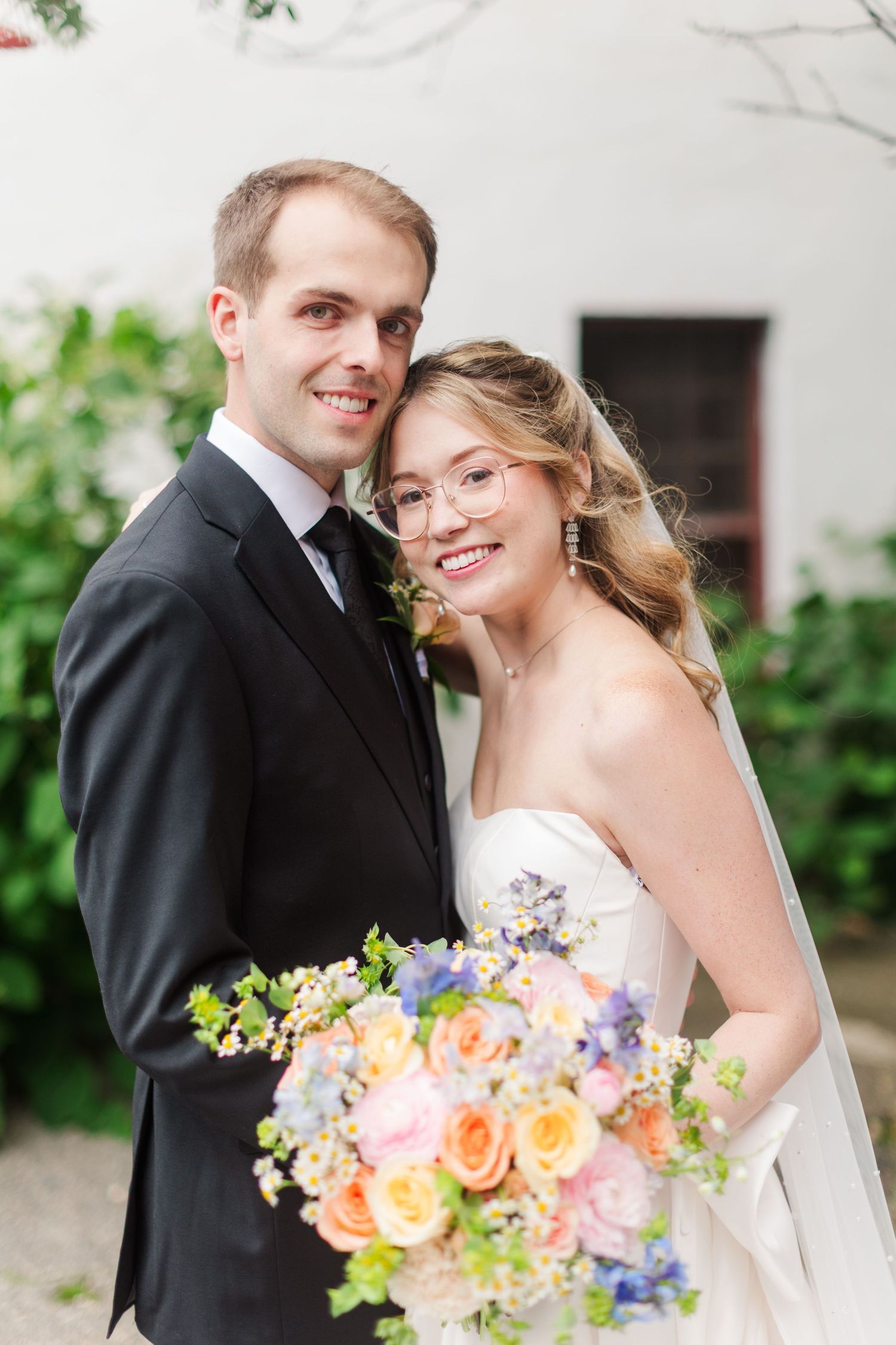 Smiling bride and groom pose together outdoors, bride holding colorful bouquet of pink, peach, and blue flowers.
