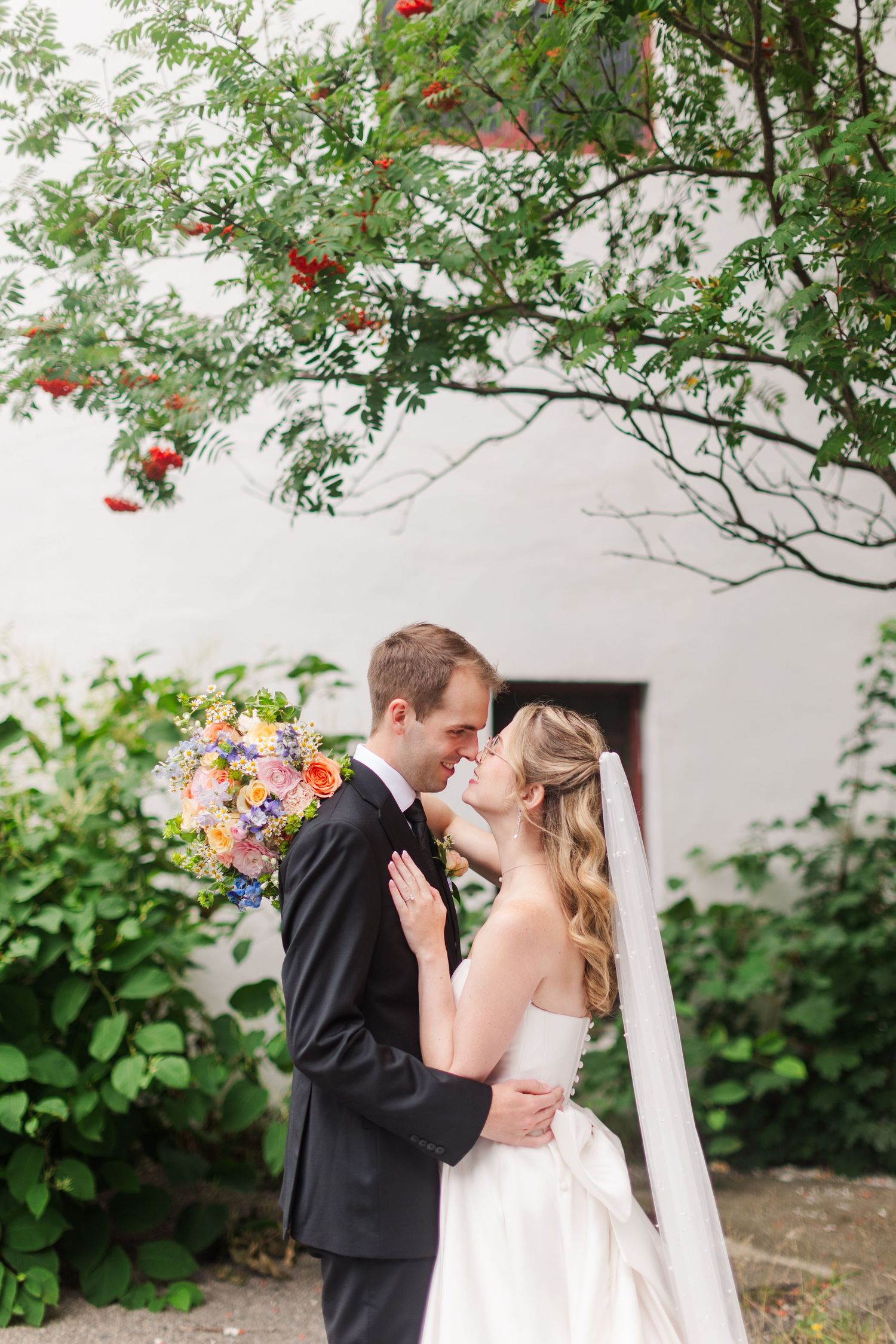 A bride and groom share a romantic moment outdoors, surrounded by lush greenery, as she holds a colorful wedding bouquet.